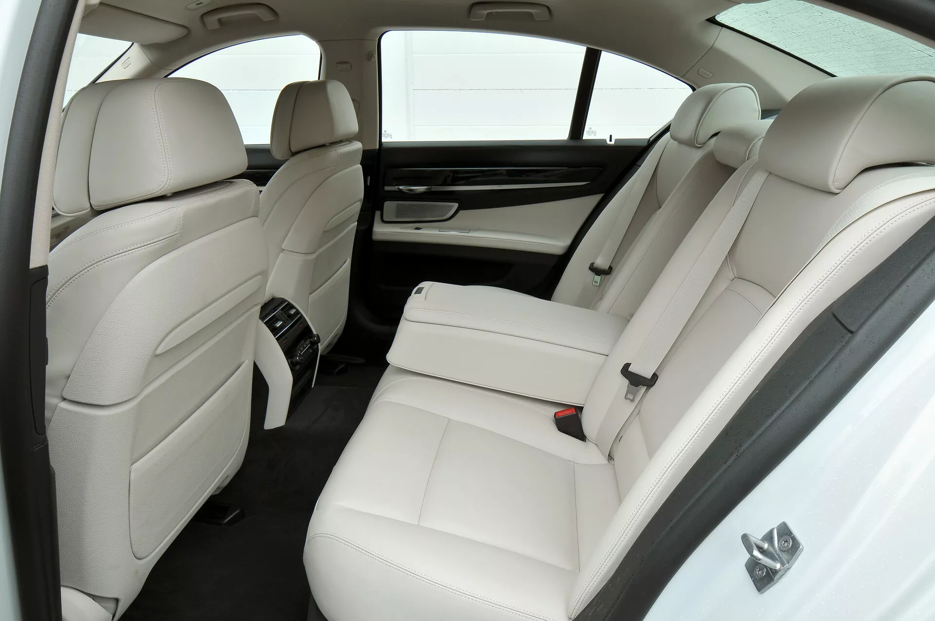 Bilden visar den lyxiga vita läderinteriören i en BMW 7-serie (2012 facelift, F01-F02) med fokus på baksätet från en något upphöjd vinkel. Den har två separata baksäten med nackstöd och sidostöd, en mittkonsol med kontroller, bakre klimatanläggningsventiler, solskydd vid bakfönstret, integrerade nackstöd på framsätenas ryggstöd och en minimalistisk färgpalett i vitt och mörka toner.