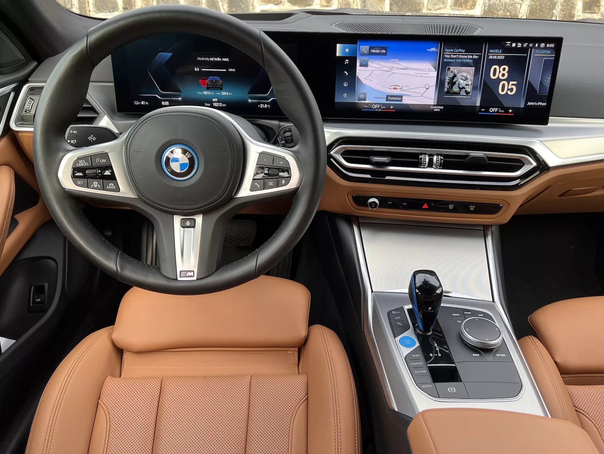 Interiörvy av en BMW i4 2021, som visar en central ratt med BMW-logotyp, multifunktionsknappar, digital instrumentpanel, pekkänsligt centralt infotainmentsystem som visar navigation och media, lyxiga trädetaljer på instrumentbrädan, klimatkontrollsgränssnitt, elektronisk växelväljare, iDrive-styrare och säten i beige läder.