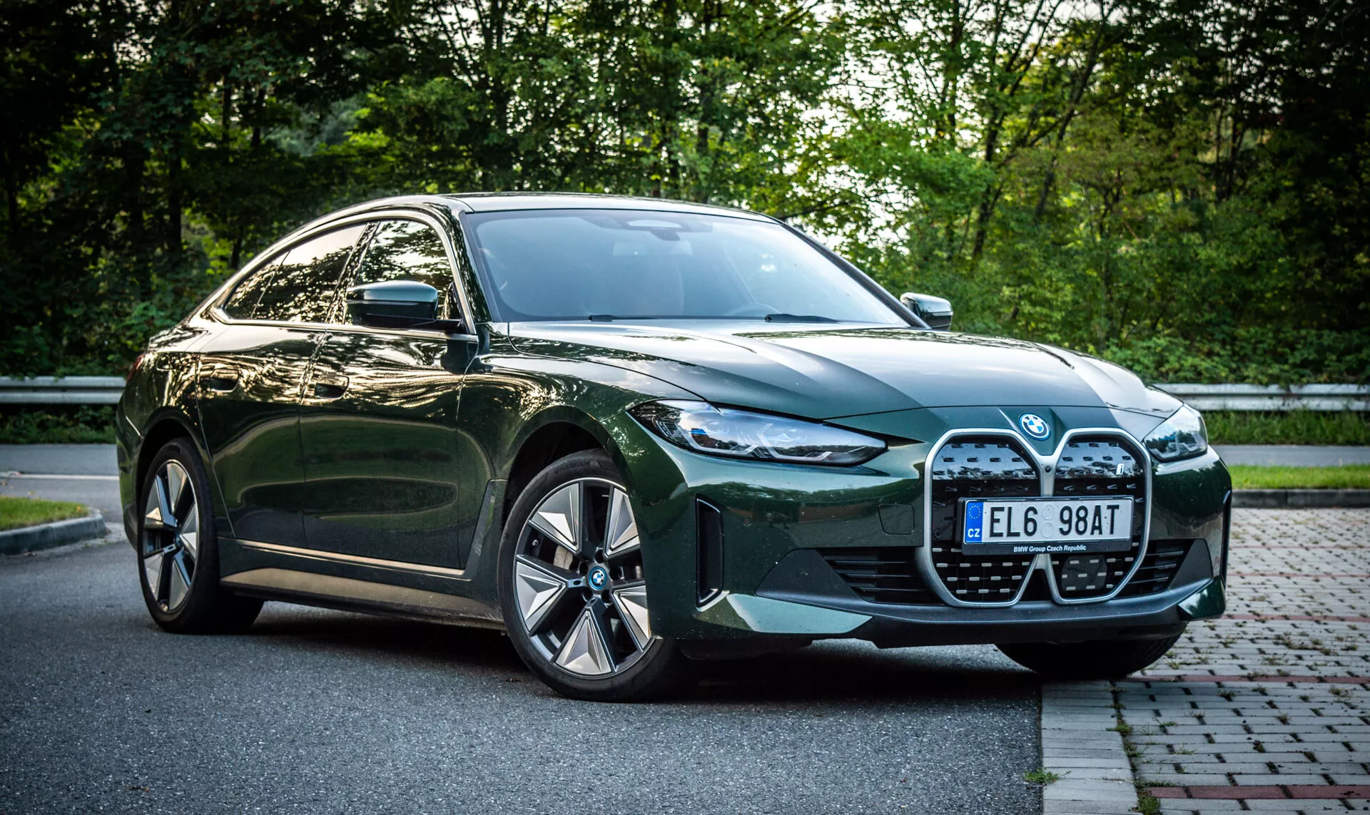 2021 BMW i4 grön fyrdörrars sedan parkerad på en gatstenbelagd gata intill ett gräsmatta med lövverk i bakgrunden, framifrån sidovy som framhäver bilens stora grill, LED-strålkastare, luftintag och legeringshjul