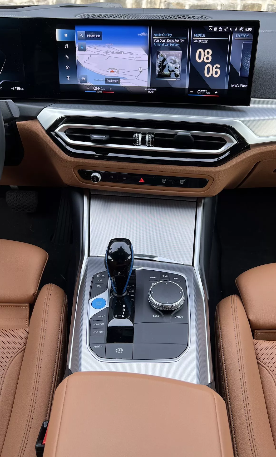 Interiör mittkonsol vy av en BMW i4 2021 från baksätena, belyser lädersätena, elektronisk växelväljare, multimedia kontrollknopp, mugghållare, klimatkontroller och dubbelskärms infotainmentskärm med navigering och media visningar