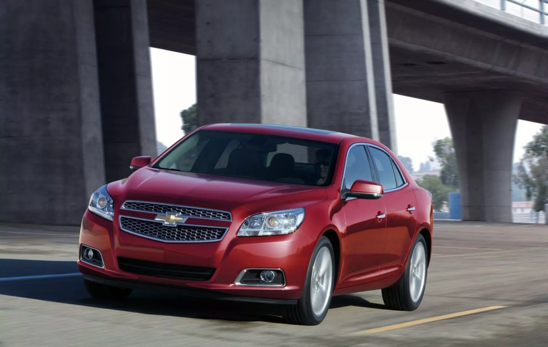 Dynamisk vy av en röd Chevrolet Malibu sedan från 2012 i rörelse på en bro, som visar fram design med kromgrill, eleganta strålkastare, sportig silhuett och legeringshjul.