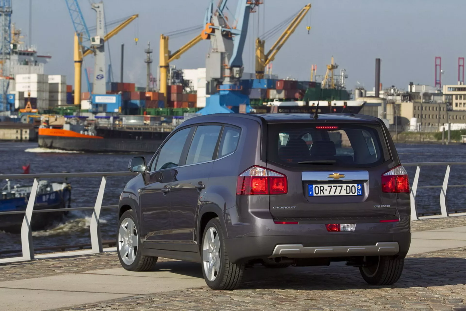 Bakifrån treskvarts vy av en Chevrolet Orlando 2011 vid en kaj vid vattnet, vilket framhäver dess kompakta SUV-kaross, femdörrarskonfiguration, dubbla avgassystem, parkeringsassistanssensorer på stötfångaren och flerspakiga aluminiumfälgar