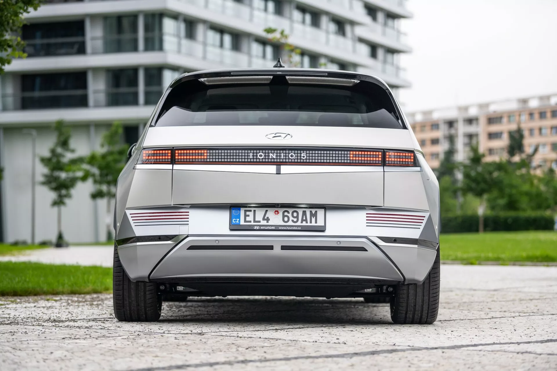 Bakåtsikt av Hyundai Ioniq 5 2WD 2021, som framhäver en distinkt bakre ljuslist över hela bredden, centralt placerad tillverkningslogotyp, platt bakdel med skarpa horisontella linjer, minimalistisk bakre stötfångare med reflekterande remsor och europeisk stild nummerplåt. Fordonet är placerat i mitten av en asfaltsyta med lövverk och moderna byggnader i bakgrunden, under molnigt ljus.