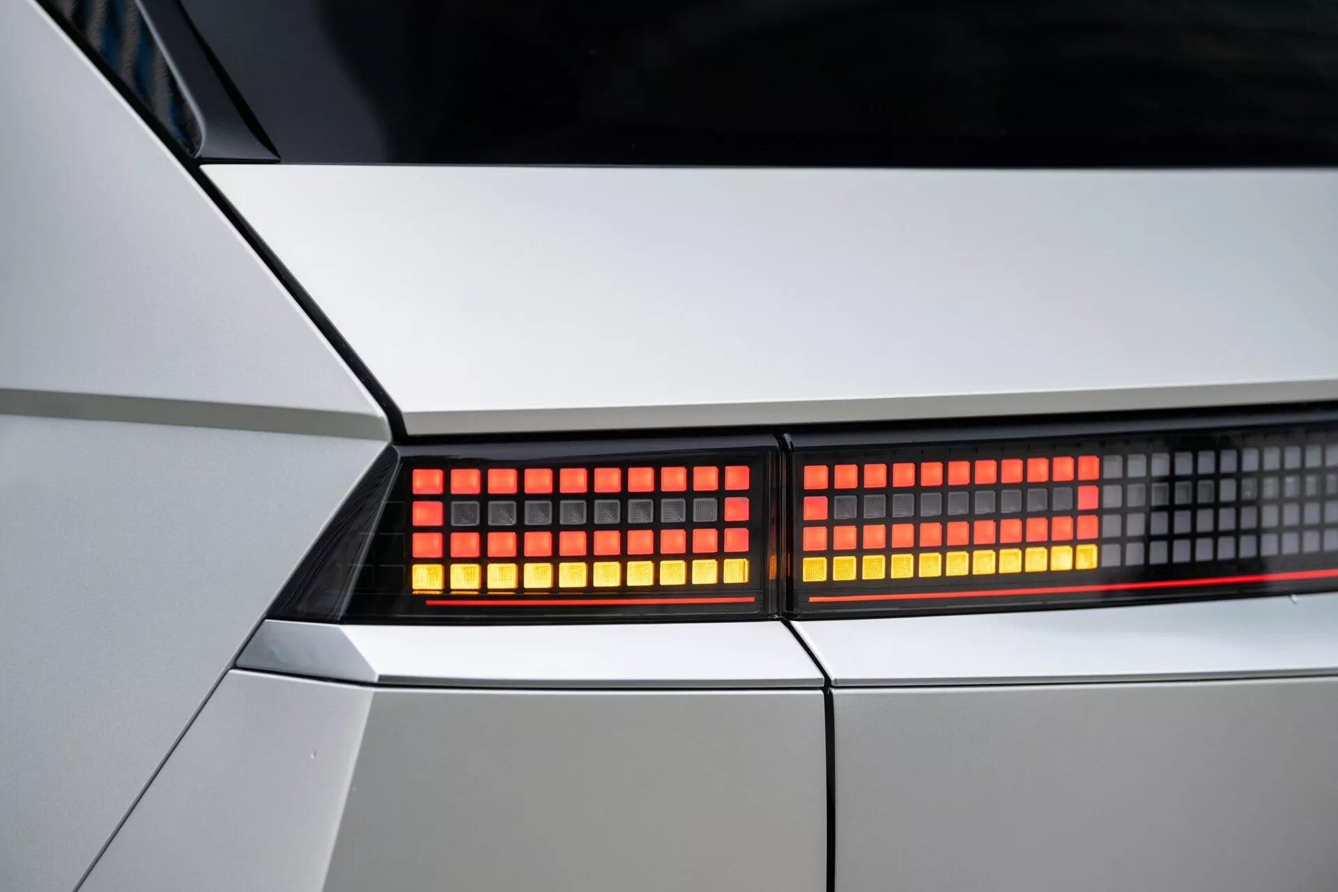 Närbild av bakre belysningsenheten på Hyundai Ioniq-5 2021 2WD. Fokuserad bild på röda, pixelbaserade LED-lampor i en snäv beskärning, som framhäver bilens skarpa, geometriska kaross i vit eller ljus silverfärg. Detaljerad vy av moderna, futuristiska designelement och belysningsteknologi.