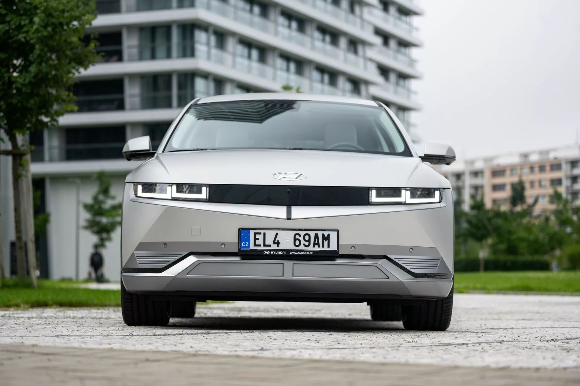 Bilden visar en Hyundai Ioniq 5 2WD från 2021, uppvisande dess framsida från en låg vinkel. Fotografiet fångar den moderna grillen, avancerade LED-strålkastare och strömlinjeformade stötfångare med detaljerade urtag, vilket betonar den aerodynamiska designen. Bilen, en elektrisk modell, har en sluten grill typisk för elfordon, en ljusstrimla under huven och sensorer för avancerade förarassistanssystem. Huvan, prydd med tillverkarens emblem, är ren och slät. Den urbana bakgrunden lyfter fram bilens lämplighet för stadsliv.