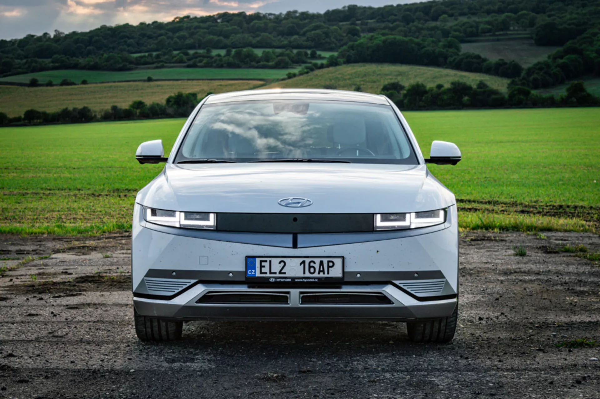 Frontvy av Hyundai Ioniq 5 elektrisk bil från 2021 på grus mot frodig grönt fält, med smala LED-strålkastare och aerodynamisk design utan traditionell grill.
