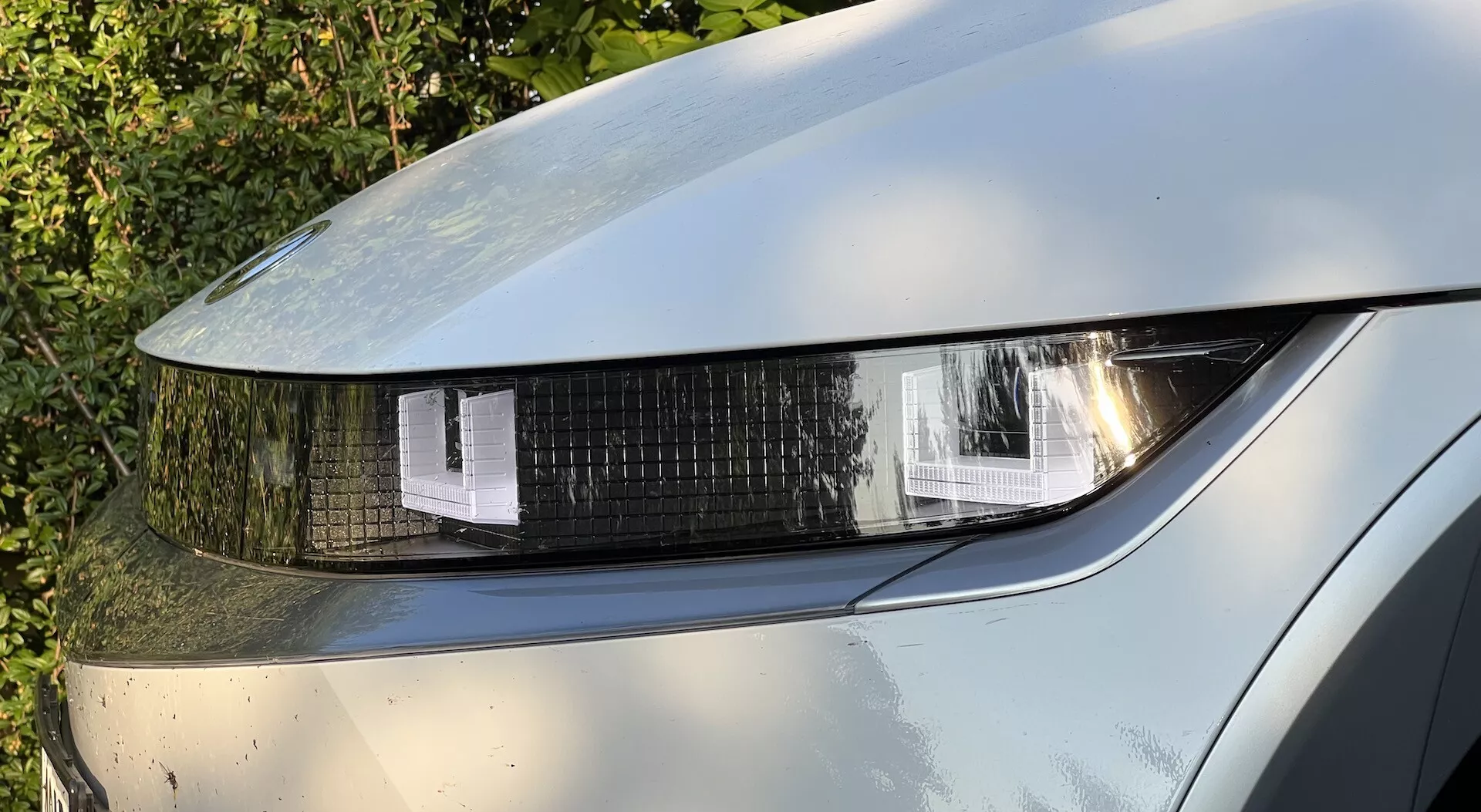 Närbild av en Hyundai Ioniq-5 2021 front vänster strålkastare, som visar LED eller HID belysning med geometriska formade segment, en glänsande klar skydd och reflektioner på metallisk målfärg med synlig smuts, vilket indikerar användning.