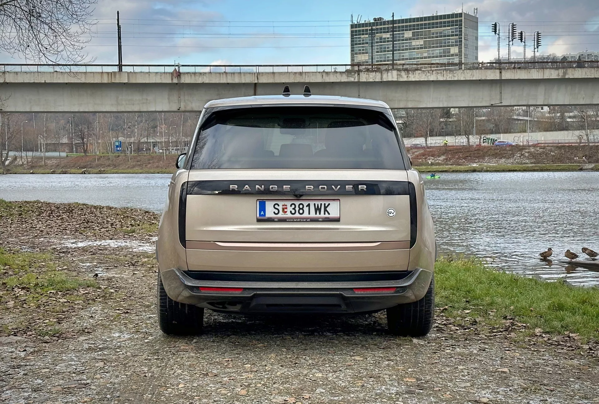 Bakvy av Range Rover 2021 med direkt ögonnivåvinkel, parkerad nära en vattenkropp med ankor och en viadukt i bakgrunden, med modellnamn, bakljus, parkeringssensorer och ett dubbelutblåssystem i matt beige
