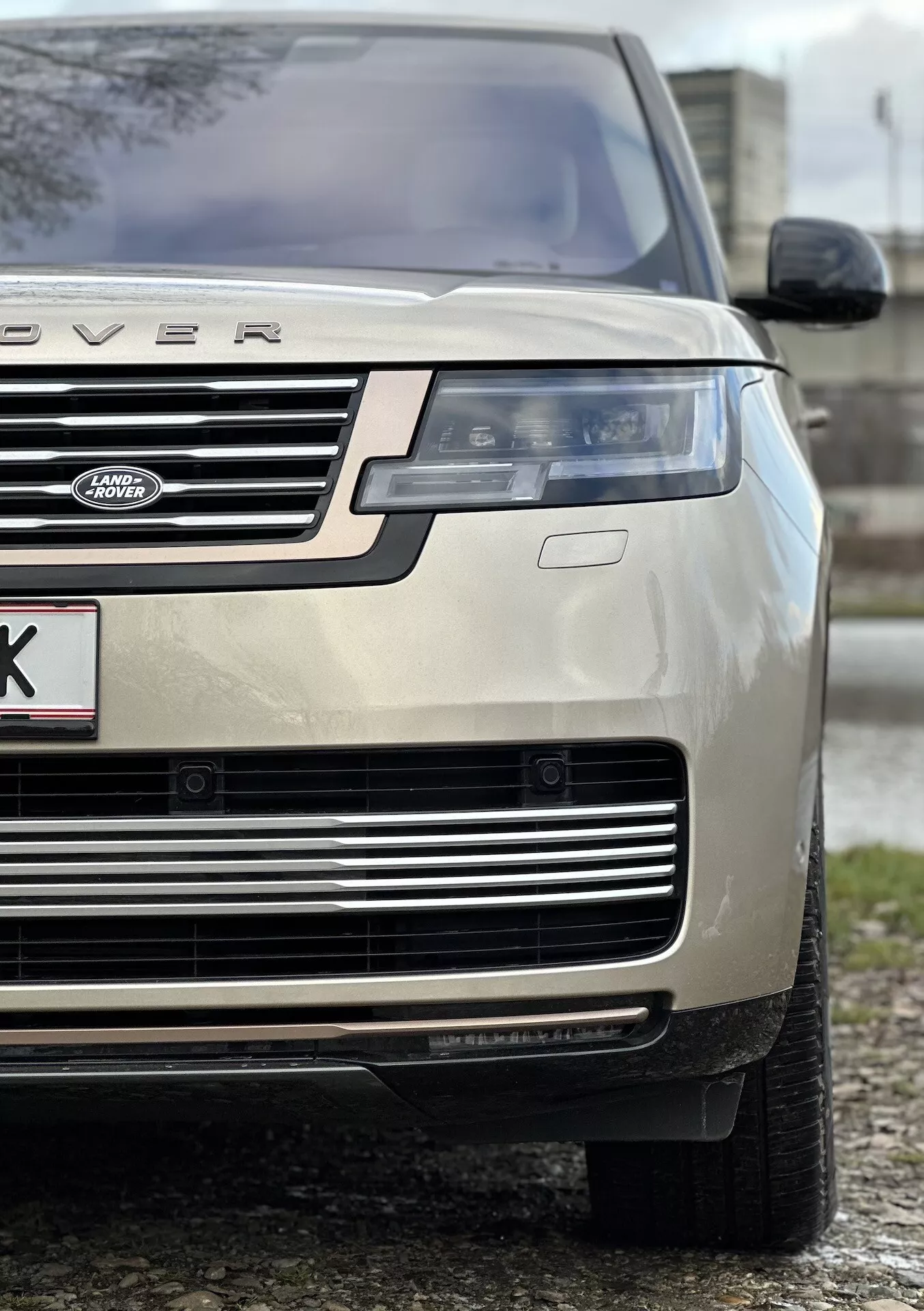 Närbild av en guld-färgad 2021 Land Rover Range Rover som framhäver fordonets främre vänstra kvartal, betonar bilens höjd och ställning med fokus på motorhuven, grillen, LED-strålkastaren, stötfångaren och allvädersdäcket, vilket indikerar lyx och terränggående förmåga.