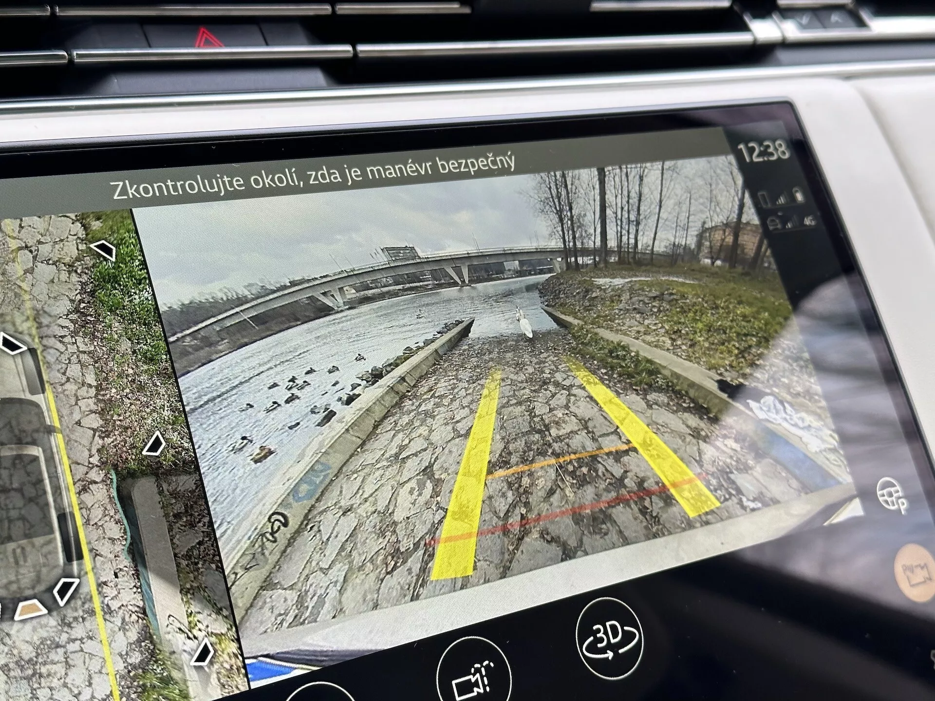 2021 Land Rover Range Rover's infotainmentskärm som visar bakvy från backkamera med dynamiska vägledningslinjer på en parkeringsplats nära en vattenfront, detaljerad med lövverk, parkeringsassistansikoner och säkerhetspåminnelsetext