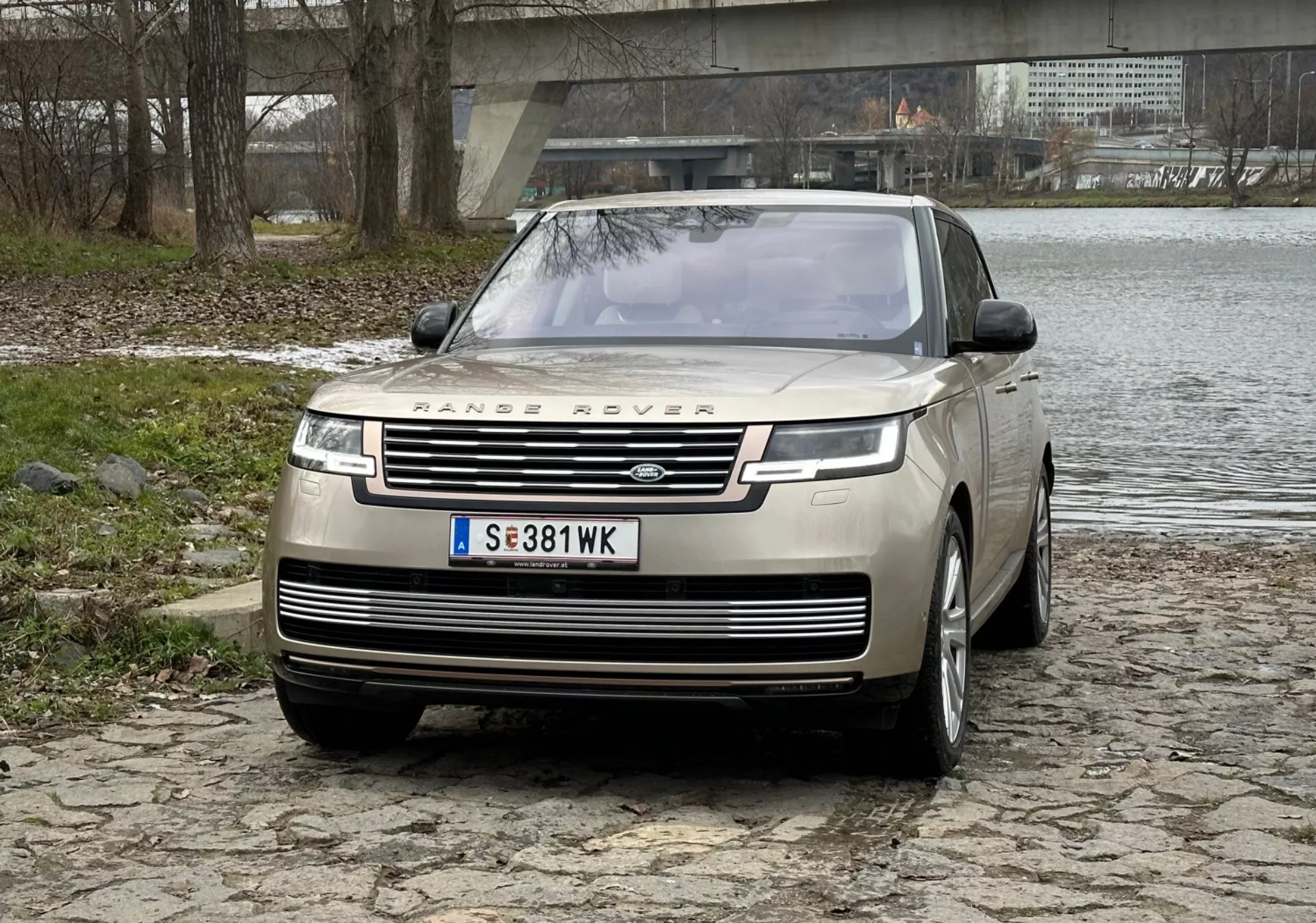 2021 Range Rover lyxig SUV i silver, visar upp framträdande frontgrill med märkesnamn, eleganta LED-dagsljus, frontstötfångare med parkeringssensorer och clamshell huva design på kullersten nära en flod