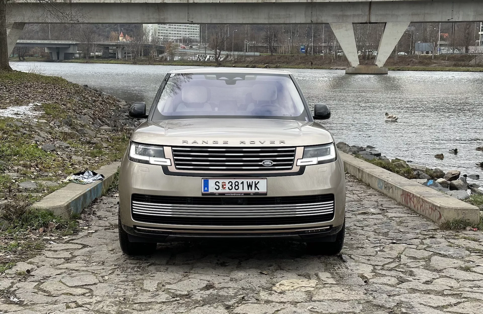 Land Rover Range Rover 2021 avbildad från en främre, något låg vinkel som betonar grillen, frontstötfångaren och motorhuven, med den ikoniska RANGE ROVER-emblemet, moderna LED-strålkastare och avancerad förarassistans teknik, mot en bakgrund av en flod- eller sjömiljö, en bro och en mulen himmel