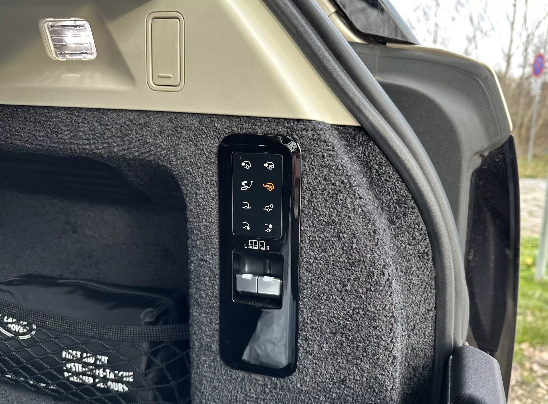 Närbild av den elektroniska bagageutrymmets kontrollpanel i interiören av en Land Rover Range Rover 2021, som visar knappar för operationer som elektronisk öppning och stängning, kontroll av intagbart lastskydd, möjligen ett bagageutrymmesljus eller hjälpkraftuttag, justeringar av bakre säten från bagageutrymmet och nödutlösning, satt mot bagageutrymmets mattvägg och ljusa tak med lastljusarmatur.
