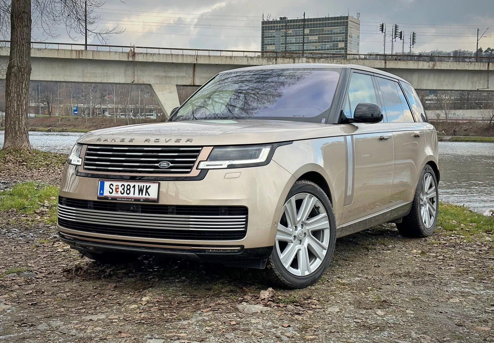 Lyxig SUV Land Rover Range Rover 2021 parkerad på blandad jord och gräsyta med en betongstruktur och vegetation i bakgrunden, visar bild från främre kvartalsvinkeln som framhäver LED-strålkastare, legeringshjul och moderna aerodynamiska egenskaper