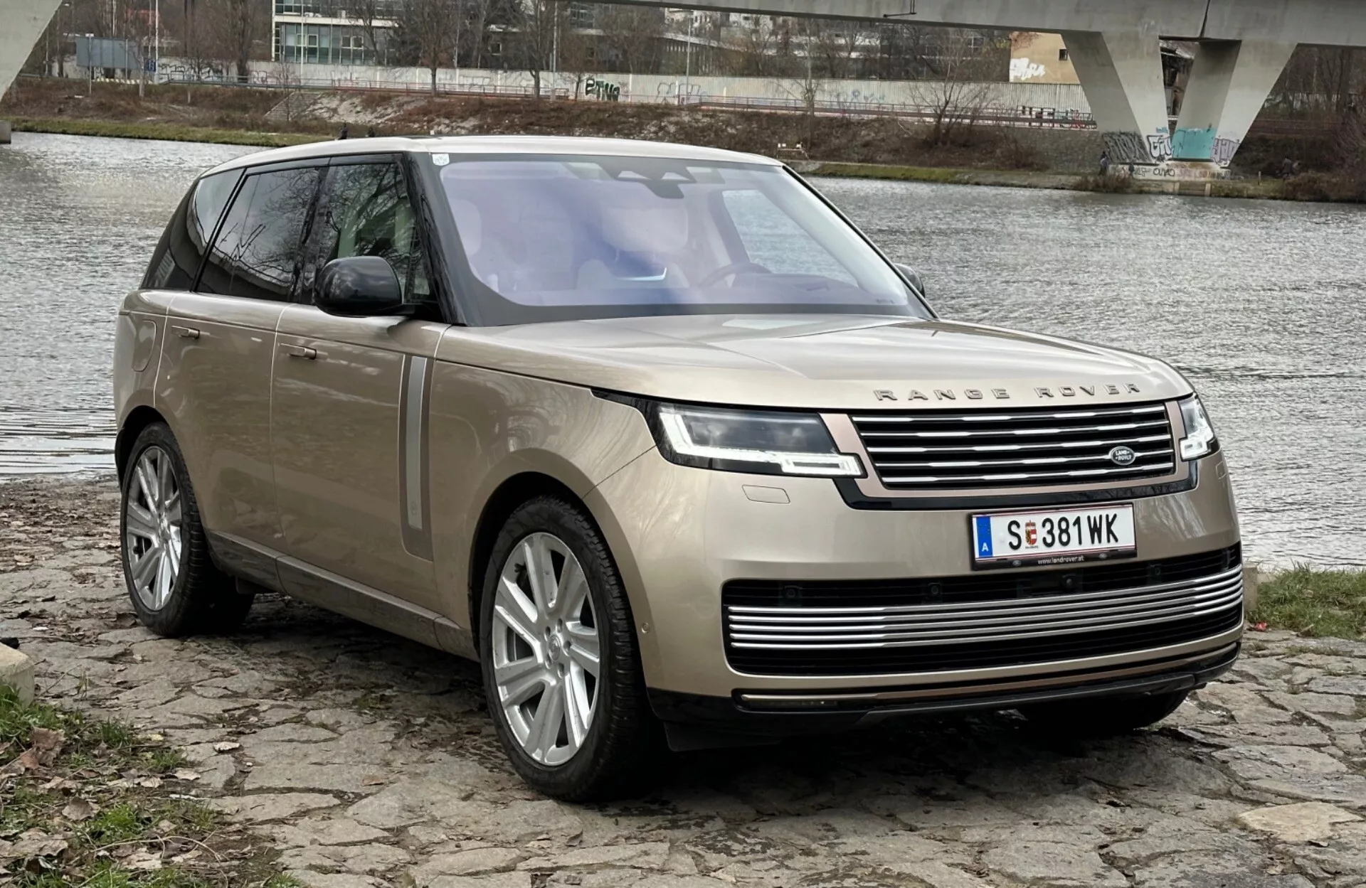 Lyxig SUV Land Rover Range Rover från 2021 parkerad vid en flodbank med en bro i bakgrunden, som visar upp sin moderna design och tekniska funktioner inklusive LED-strålkastare, frontgrill med emblem, flerspakiga legeringshjul och panoramatak.