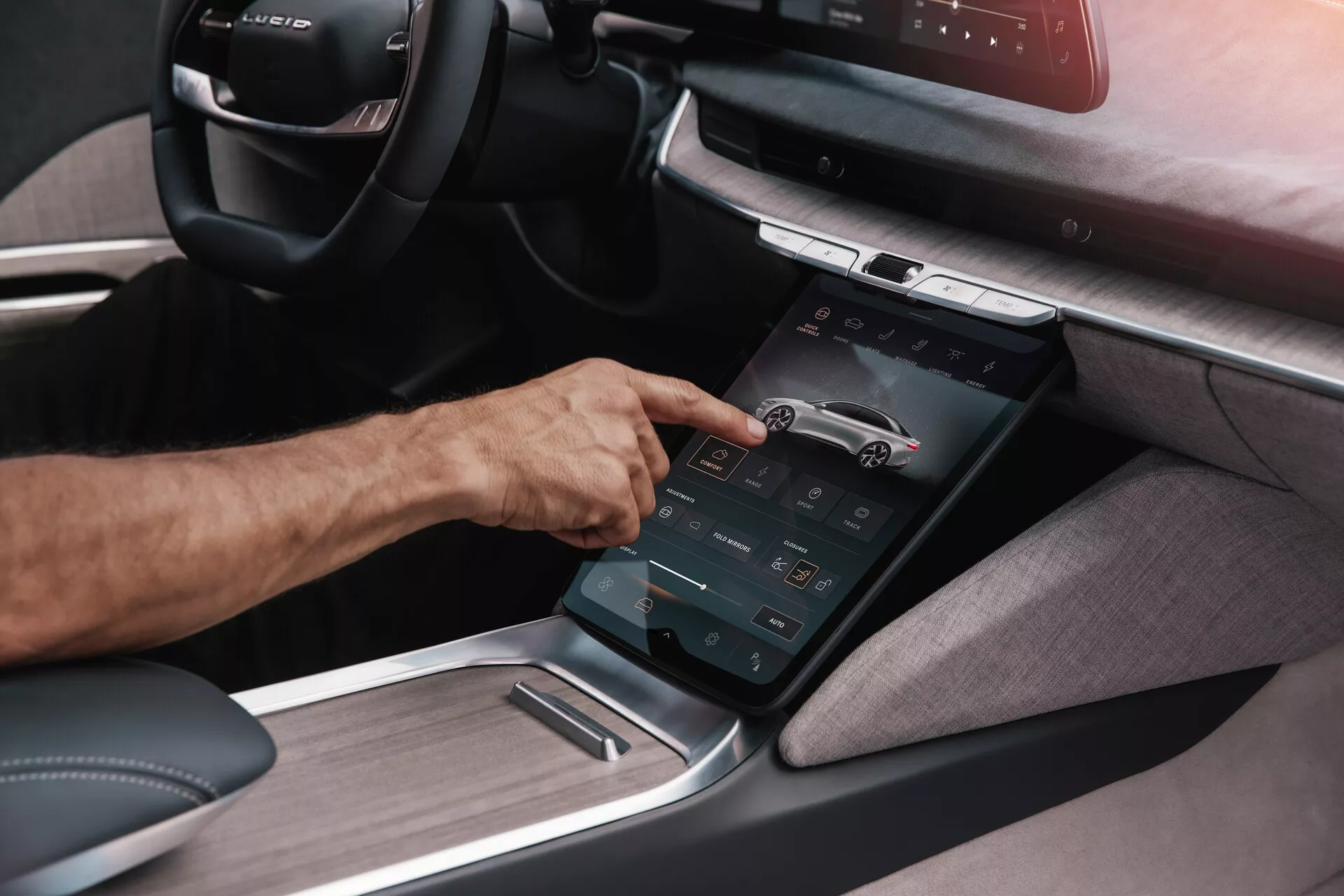 Detaljerad vy av centrokonsolen i Lucid Air 2022 av första generationen från en sned vinkel, med betoning på en vertikalt orienterad pekskärm som interagerar med en persons högra hand, som illustrerar det avancerade infotainmentsystemet med olika funktionskontroller och en kombination av textil- och metallpaneler, vilket tyder på en modern, lyxig interiördesign med fokus på högteknologi.