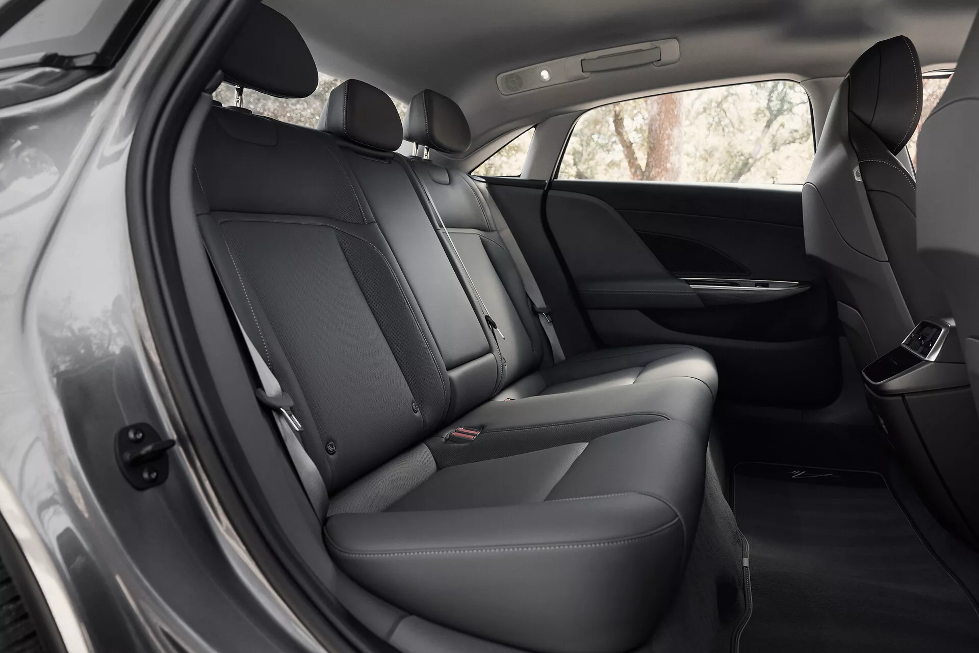Interiörvy av en Lucid Air 2022 första generationen, fokuserad på det bakre sittområdet från en något framåtvänd vinkel. Sätena har mörkt läder eller premium syntetiskt klädsel med synliga sömmar. Dörrpanelen visar en högtalare, elektrisk fönsterstyrning och möjliga förvaringsfack. Framsätenas ryggstöd och hytten är rena och väl underhållna, belysta av naturligt ljus.