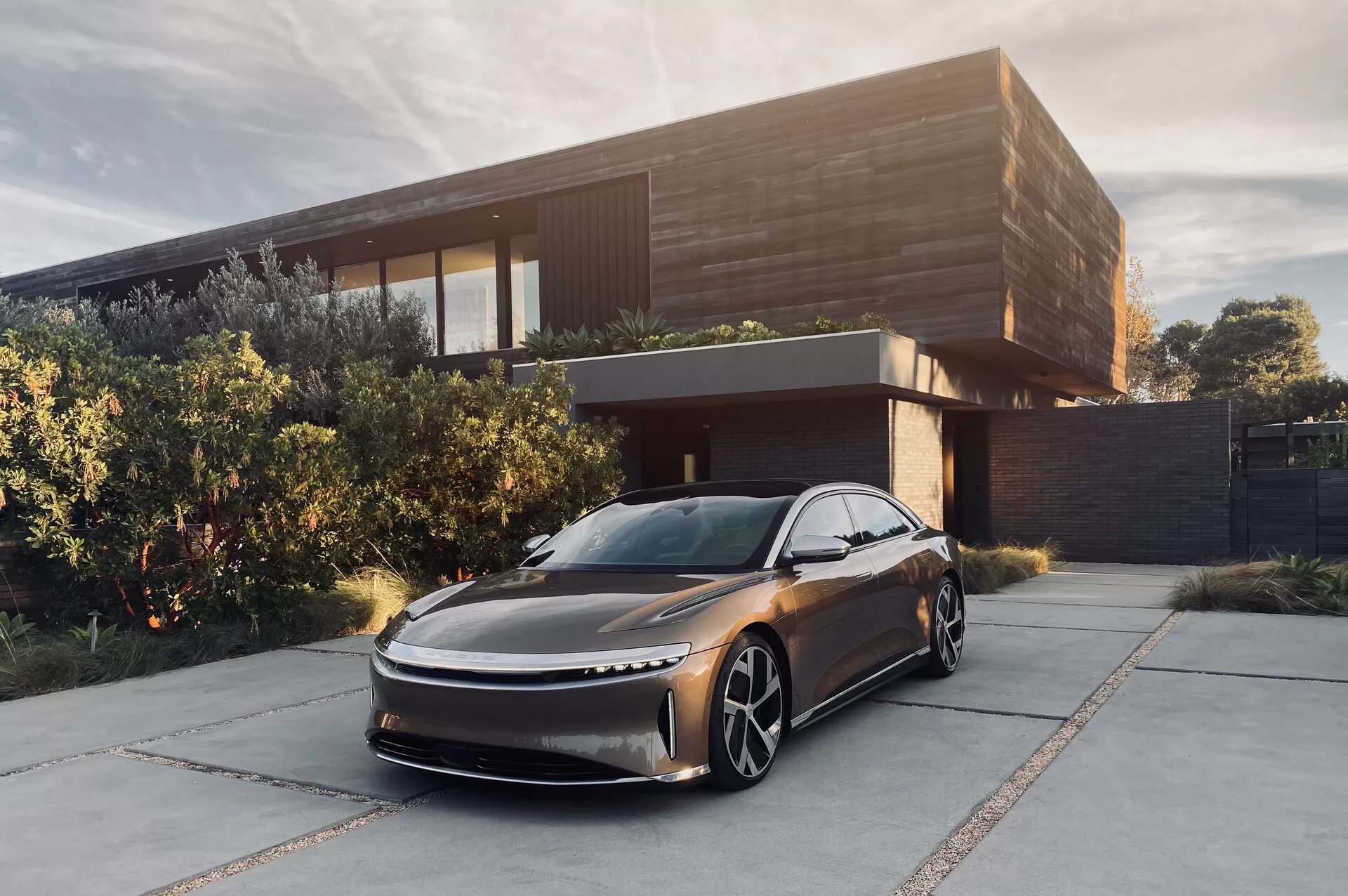 Bild av 2022 Lucid Air, en modern sedan, parkerad framför ett samtida hus. Fotograferad från en låg, trekvartsvinklig vinkel som visar upp dess eleganta, aerodynamiska design med en förlängd huv och subtilt gallerområde. Framträdande detaljer inkluderar tunna LED-strålkastare, stora distinkta hjul och inga synliga sidospeglar, vilket hintar om kamerabaserade system för bakåtsikt. Den mörka metallic-färgen och den sofistikerade miljön förstärker dess lyxiga känsla, vilket antyder att det är ett elfordon riktat mot en förmögen marknad.