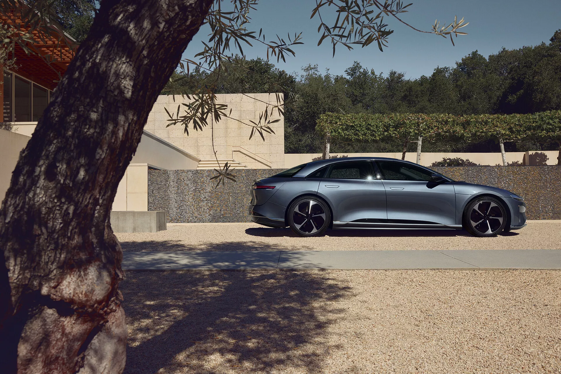 Profilvy av en fyrdörrars sedan Lucid Air 2022 första generationen parkerad utomhus. Passagerarsidan av den lyxiga, prestandainriktade sedanen är synlig, vilket visar upp de släta karosslinjerna, designen av dörrar och fönster samt de distinkta legeringsfälgarna med lågprofildäck. Delvis skuggad av trädlöv, med naturligt ljus som framhäver den metalliska finishen. En modern byggnad i bakgrunden framhäver bilens eleganta, aerodynamiska design med avancerade funktioner som infällda dörrhandtag och aerodynamiska sidospeglar som möjligen antyder integrerade blinkers eller dödvinkelavkänning.