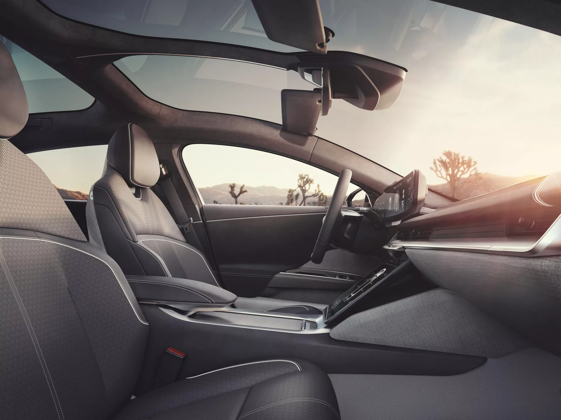 Interiörsbild av 2022 Lucid Air (1:a generationen) från baksätespassagerarens perspektiv, som visar mörka lädersäten med detaljerad sömnad, elegant instrumentpanel med vänsterstyrd ratt med integrerade kontroller, framträdande pekskärmsinfotainmentskärm, ergonomisk växelspak, dörrpaneler med elektroniska kontroller och högtalare, synlig backspegel och eventuell förarhjälpsanordning vid vindrutan, som betonar lyxig design, avancerad teknologi och komfort.