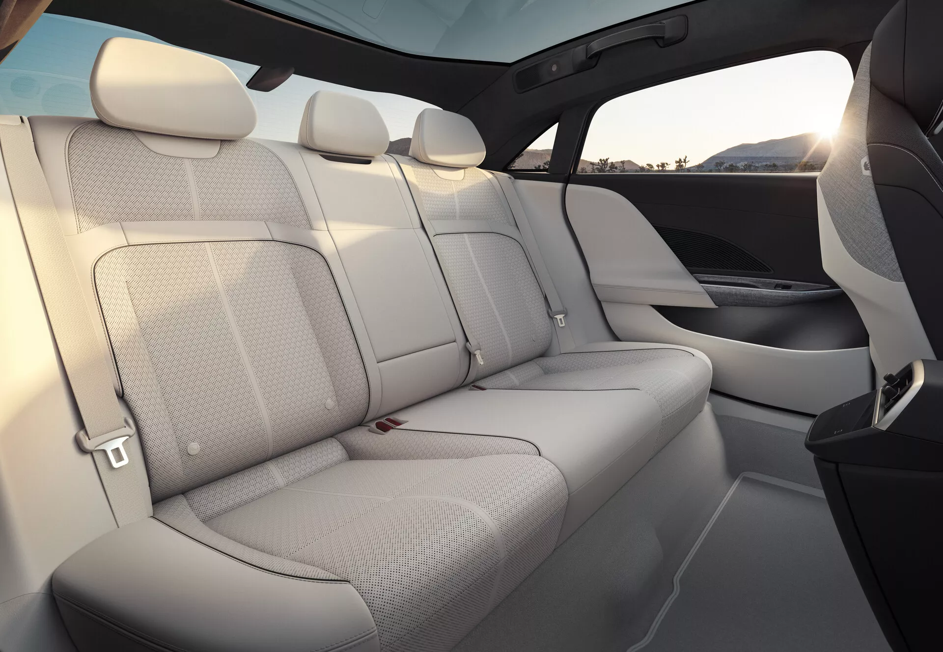 Interiörvy av Lucid Air 2022, med fokus på baksätena. Sätena är klädda i ljusfärgat läder och ger en luftigt och rymlig känsla. Den bakre bänken rymmer tre passagerare med matchande nackstöd och synliga säkerhetsbälten. Dörrpanelerna smälter sömlöst ihop med sätesmaterialet och designen och har inbyggda högtalargaller. En hög fönsterlinje och ett expansivt tonat panoramasoltak förstärker känslan av avskildhet och skydd. Inredningen visar en blandning av ljus klädsel och mörkare element, sannolikt av högkvalitativa material.