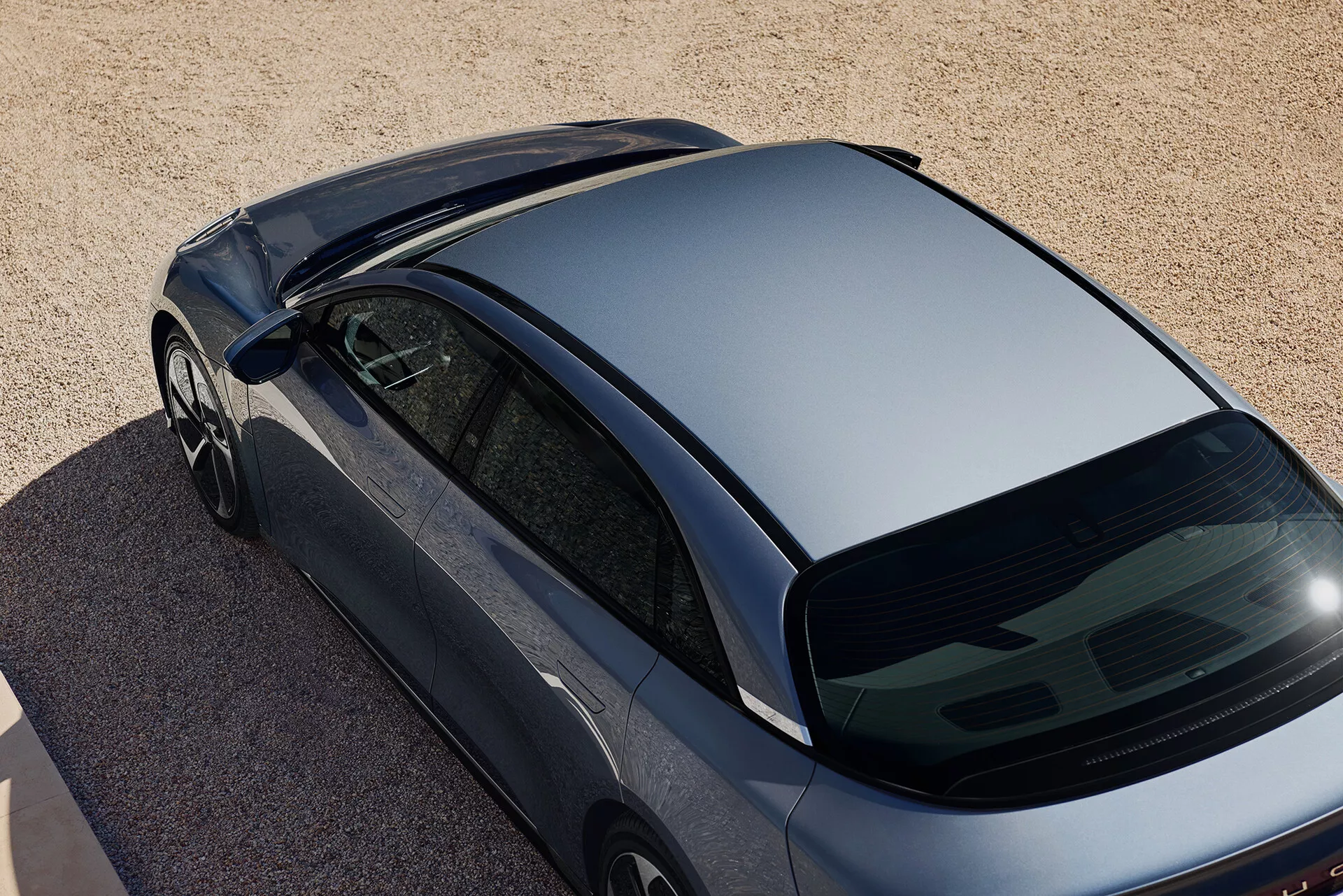 Överliggande vinklad bild av en mörk metallisk Lucid Air 2022 första generationen, parkerad på en yta av små stenar eller grov sand. Bilden fångar top-down-vyn med fokus på det strömlinjeformade taket, den brant lutande bakrutan och en del av bagageutrymmet. Funktioner inkluderar ramlösa fönster, strömlinjeformade sidospeglar och frånvaron av taktillbehör och bakrutetorkare, vilket tyder på aerodynamisk design och modern styling.