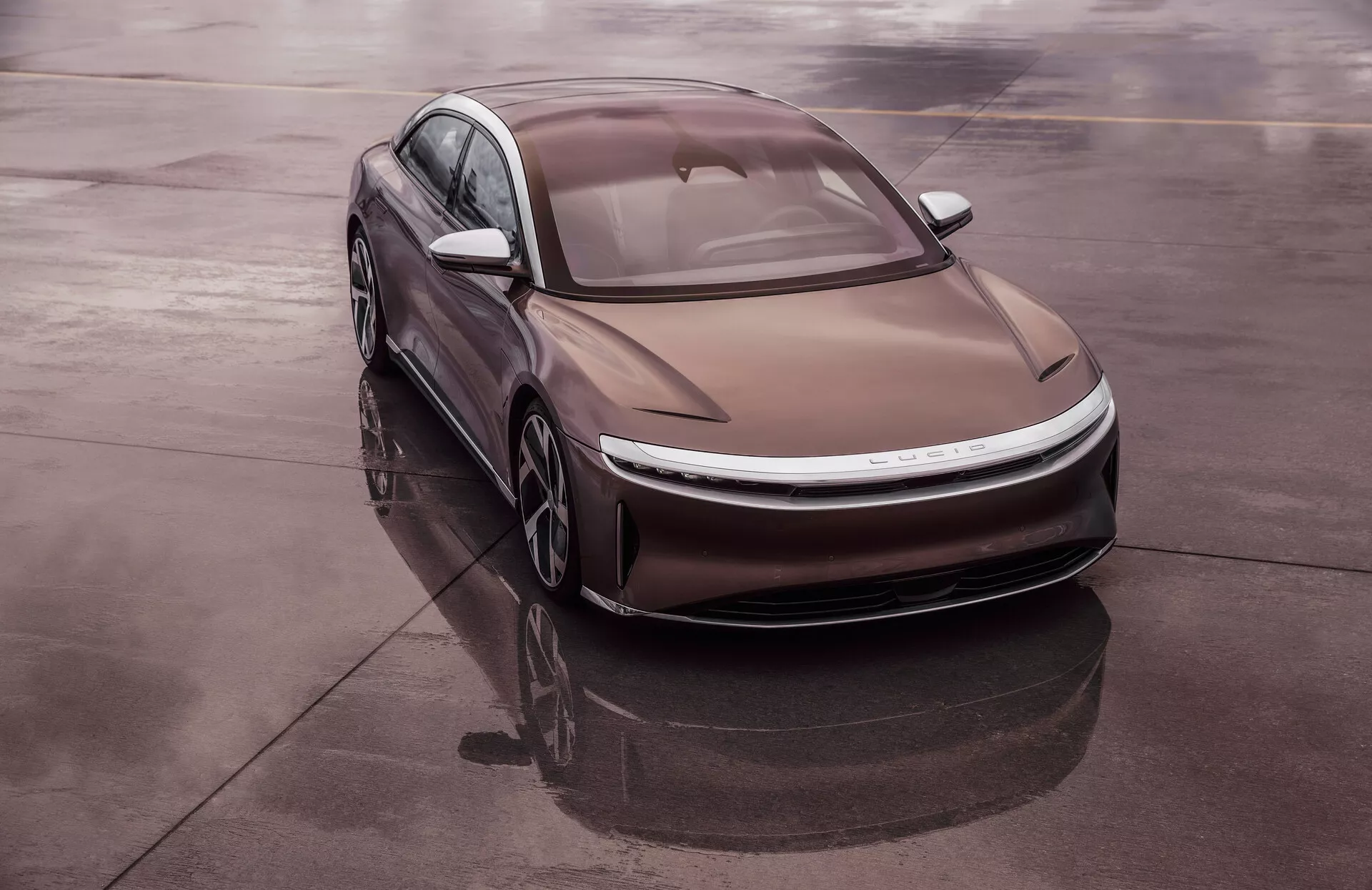 En högvinklad bild av den moderna sedanen Lucid Air 2022 från första generationen, diagonalt placerad på en reflekterande yta som våt asfalt, vilket ger en omfattande vy av fordonets vänstra framdel. Den eleganta, aerodynamiska designen har mjuka kurvor, smala LED-strålkastare och en konturerad motorhuv med framträdande linjer. Taklinjen påminner om en fastback och glaset är jämnt med karossen, vilket förbättrar den aerodynamiska effektiviteten. Frånvaron av traditionellt galler och det integrerade luftintagssystemet antyder att det är en elbil, komplett med lätta aerodynamiska hjul.