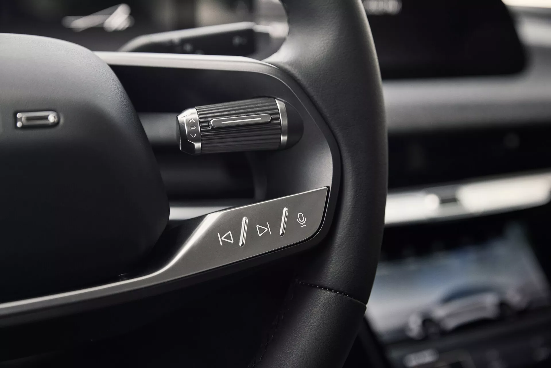 Närbild av vänstra sidan av Lucid Air 2022:s ratt som visar multifunktionella mediakontrollknappar. Fotot, taget underifrån från en sittande förares bröstnivå, framhäver det högkvalitativa lädermaterialet med metalliska accenter och en paddel bakom ratten, vilket indikerar avancerade köregenskaper.