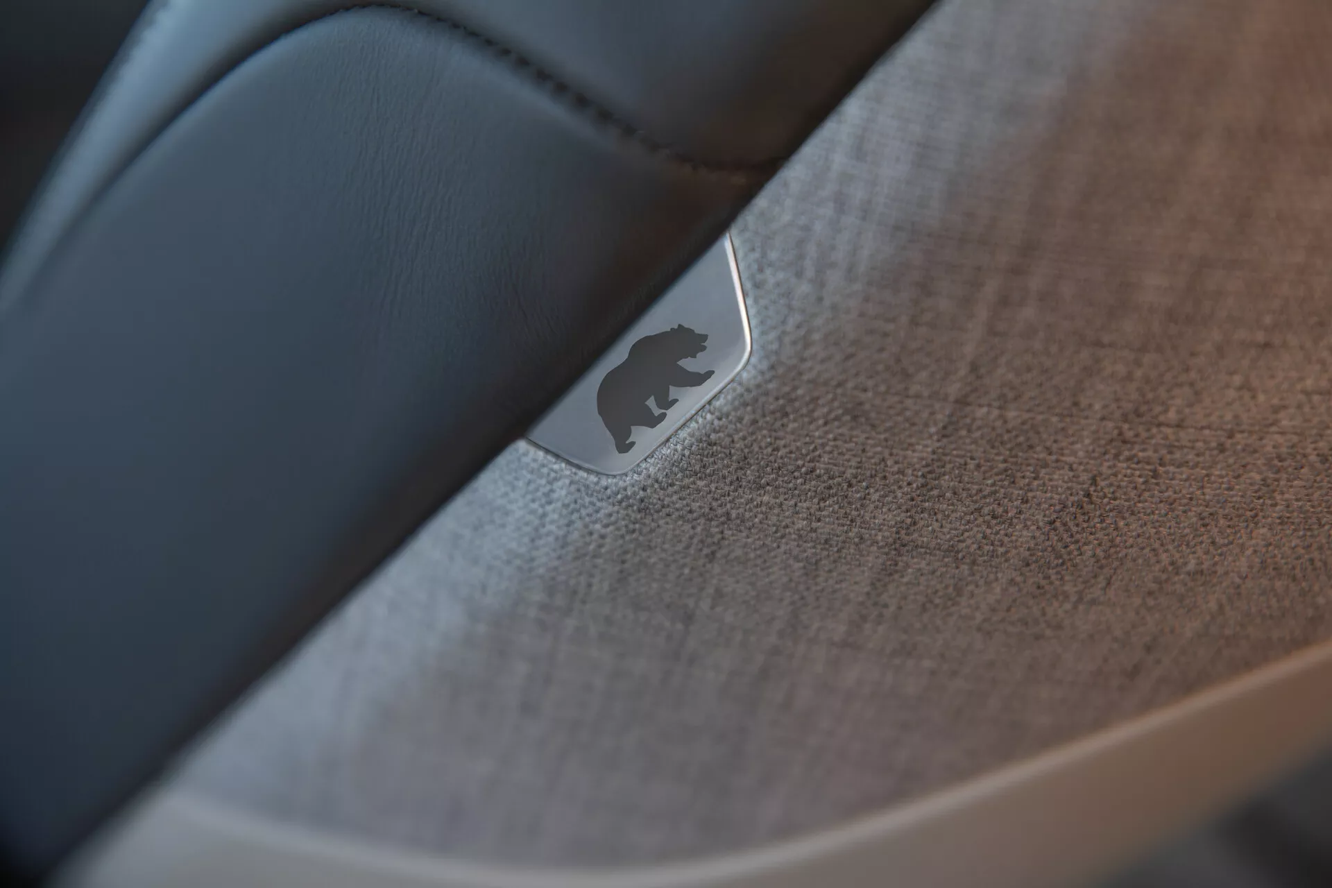 Närbild, selektivt fokuserad bild av interiören i Lucid Air 2022 (1:a generationen) som betonar detaljen på en dörr eller instrumentbräda. Ett reflekterande metallmärke eller emblem är framträdande, placerat på ett texturerat tyg eller mjuk plast med synliga sömmar. Ett slätare, mörkare material, troligtvis svart läder, skapar en kontrast mot de texturerade elementen, vilket visar på den hantverksskicklighet och höga kvaliteten i bilens interiördesign.