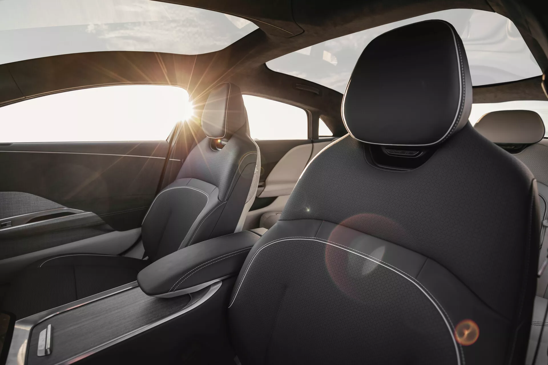 Interiörvy av Lucid Air 2022 sett från ett baksätesperspektiv med fokus på förarsätet. Bilden framhäver premium-material och design, med detaljerade sömmar på läderklädseln, ergonomiskt utformade nackstöd, borstad metallaccent och en linsreflektion från solen genom vindrutan.