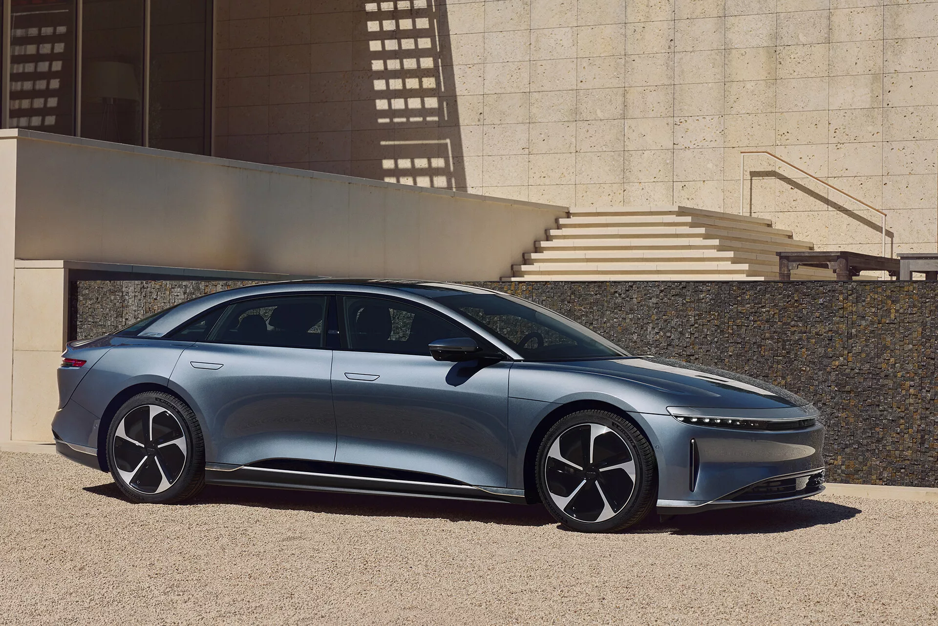 En sidoprofil av Lucid Air 2022, en modern, elegant sedan, fångad utomhus i en urban miljö med arkitektoniska element som kastar skuggor. Bilens vänstra sida visar dess aerodynamiska design med en låg taklinje, infällda dörrhandtag, stora flerspoked hjul och smala sidospeglar. Bilden framhäver bilens längd från frampanel till bakpanel, med en framåtriktad A-pelare som ger en rymlig interiörsutseende och en dynamisk karaktärslinje på karossen.
