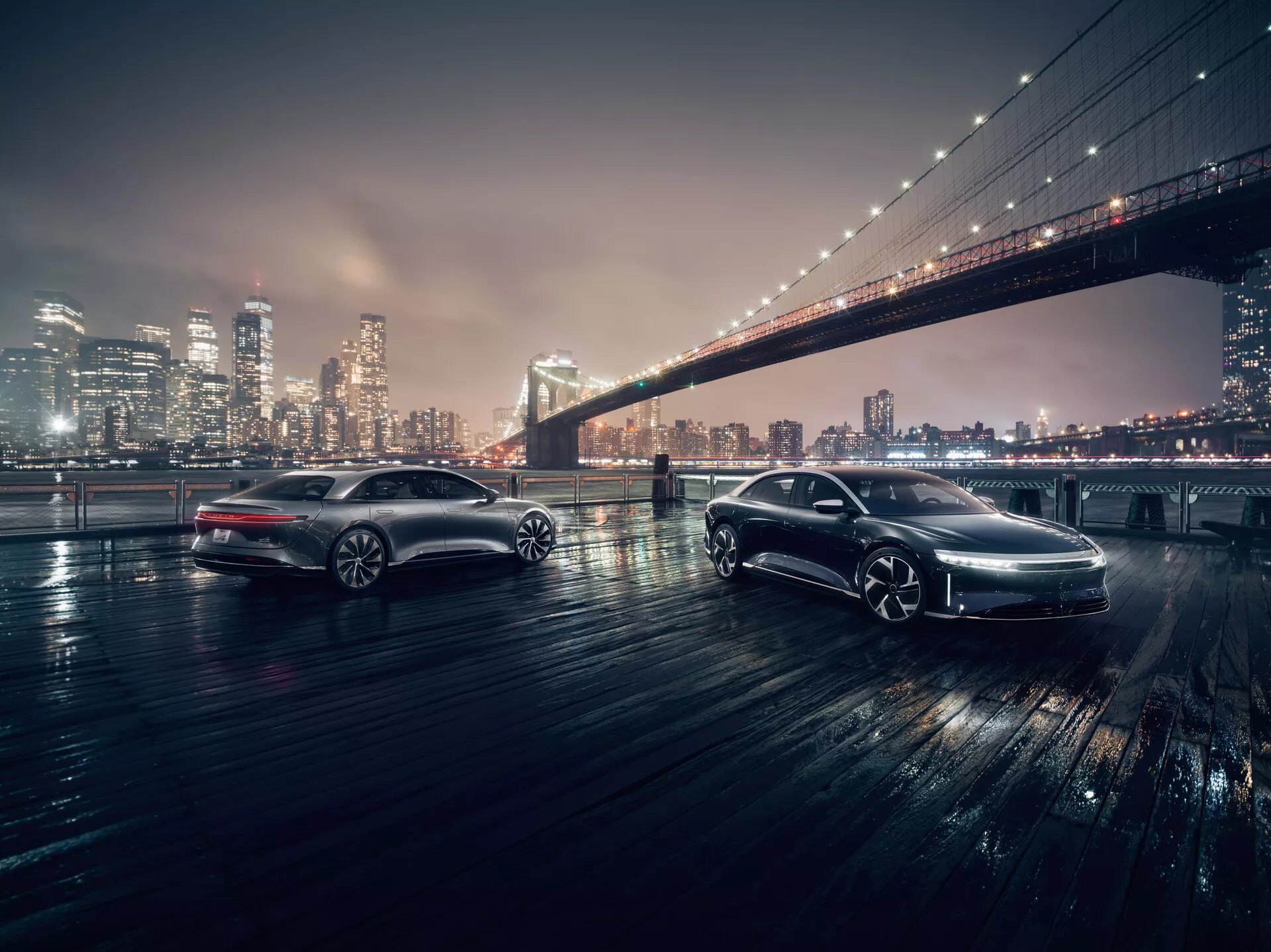 Två eleganta bilar i en nattlig stadsbild med den ikoniska Brooklyn Bridge i bakgrunden. Bilarna är fotograferade från en låg vinkel, vilket framhäver deras designdetaljer. Den främre bilen, vinklad mot kameran, visar sin grill, LED-strålkastare och regnsensor-torkare. Den bakre bilen, parallell med kameran, visar sin sidoprofil, lättmetallfälgar och högkvalitativ lack. Båda fordonen står på en våt yta som reflekterar deras LED-belysning, vilket tyder på avancerad teknik som ABS och antisladdsystem.
