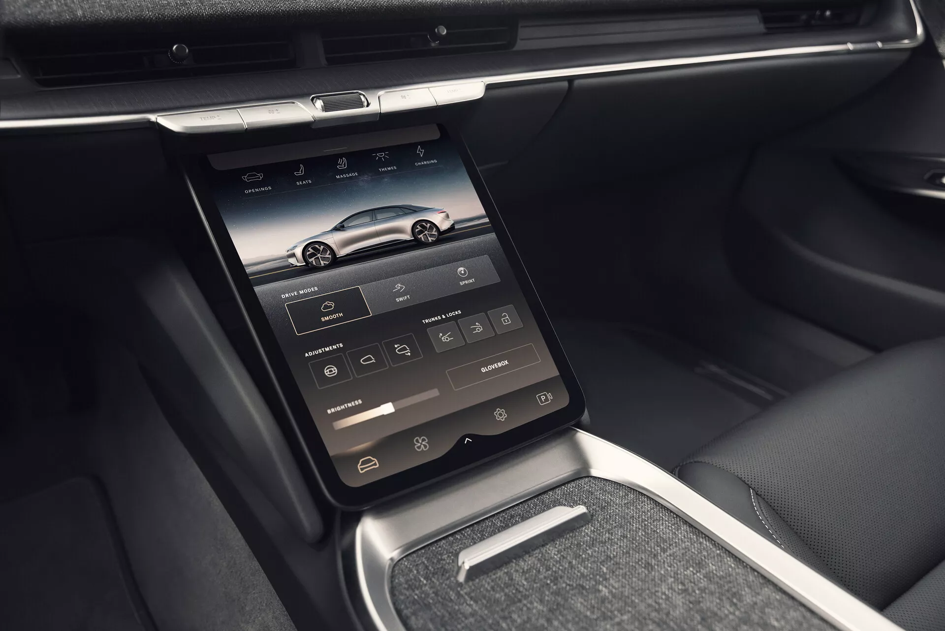 Interiörvy av Lucid Air 2022 (första generationen) som visar mittkonsolen med en vertikalt monterad integrerad pekskärm, med ikoner för navigering, telefon, media, klimatkontroll och fordonsinställningar. Fotot, taget från en något vänsterlutad vinkel, lyfter fram avancerade teknologiska funktioner med ventilationsöppningar ovanför skärmen, glänsande krom- eller metallinredning, stilren instrumentbräda och en blandning av läder- och metallfinish. Bilden detaljerar också en liten hylla under pekskärmen, en metallväxelväljare och läderklädda armstöd och säteskanter, som betonar en lyxig och bekväm interiör.