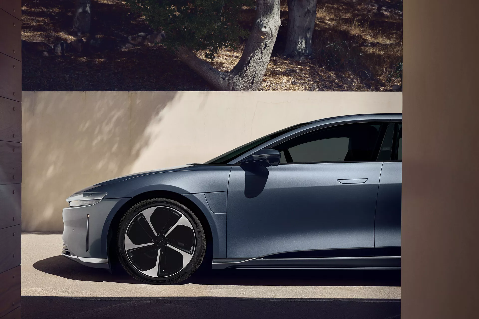 Lucid Air 2022 första generationen modern bil sidoprofilvy från en låg vinkel, vilket framhäver en stabil och kraftfull hållning. Synliga funktioner inkluderar framdörr, sidopanel, framhjul, infällbara dörrhandtag, stor aerodynamisk hjuldesign, slank minimalistisk kaross, avsaknad av traditionell grill som tyder på elektriskt drivsystem, tonade rutor, svart A-stolpe som kontrasterar mot karossfärgen, och antydan till en aerodynamisk sidospegel.