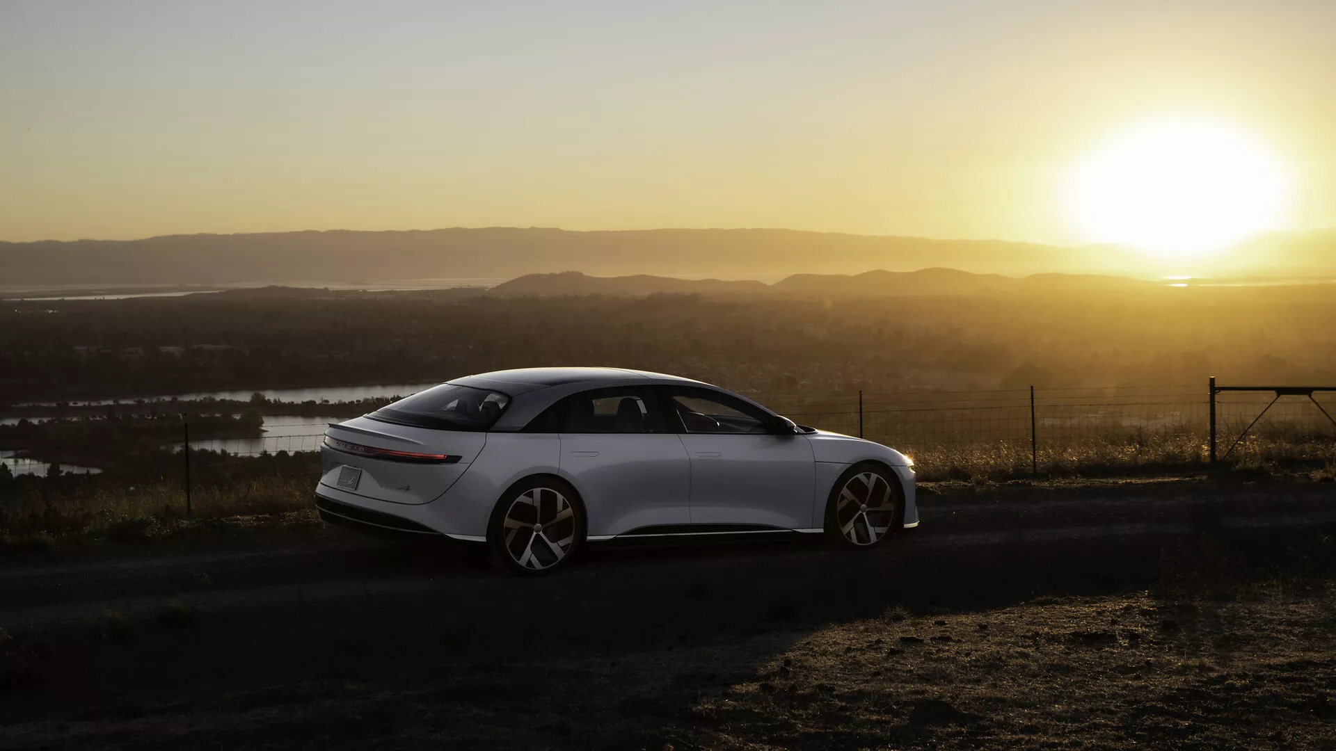 Lucid Air 2022 (1. Generationen) fotograferad från ett bakre sidovinkel under soluppgång eller solnedgång med en varm sol i bakgrunden, placerad på vänster sida av ramen. Den eleganta, moderna fyrdörrars sedanen har släta linjer, stora flerspokedesignade hjul och infällbara dörrhandtag. Landskapet inkluderar vattenkroppar och en hög utkikspunkt. Baklyktorna är smala och horisontellt förlängda, och solljuset skapar en varm lyster och en linsflare-effekt på scenen.