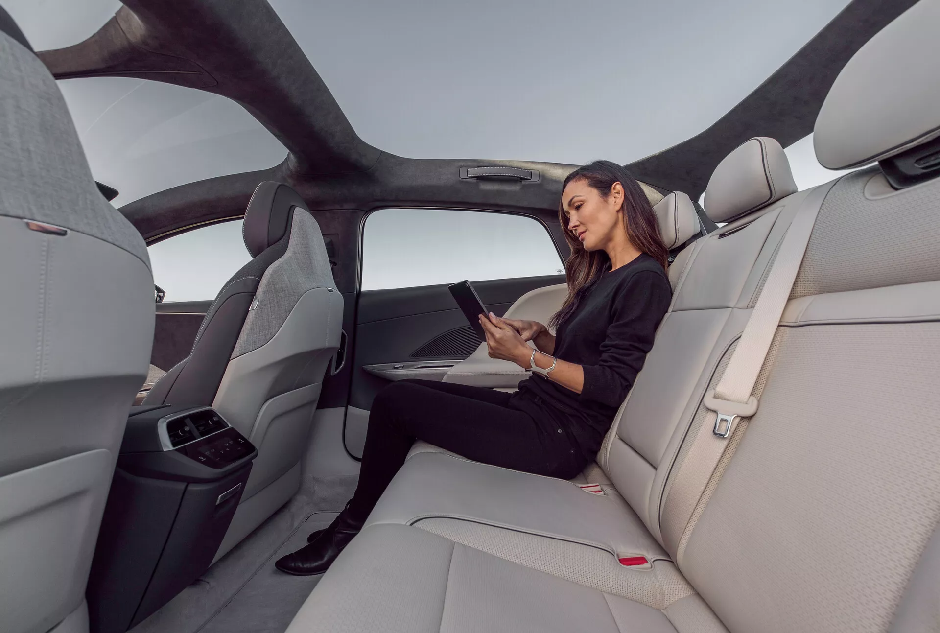 Interiörvy av baksätet i en Lucid Air 2022, som framhäver den rymliga och lyxiga kupén med ljusgrå, formade lädersäten. Bilden fångar en vidvinkelbild från höger sida, som visar panoramasoltaket som sträcker sig över det bakre utrymmet, ergonomiska sätesdesigner och en nerfälld mittarmstöd med mugghållare. En kvinna sitter bekvämt och interagerar med en surfplatta, vilket indikerar potentiella anslutningsfunktioner. Elegant, minimalistisk interiör med röda frigöringsknappar för säkerhetsbälte och horisontella linjer på instrumentbrädan och dörrpanelerna som förstärker känslan av bredd och öppenhet.