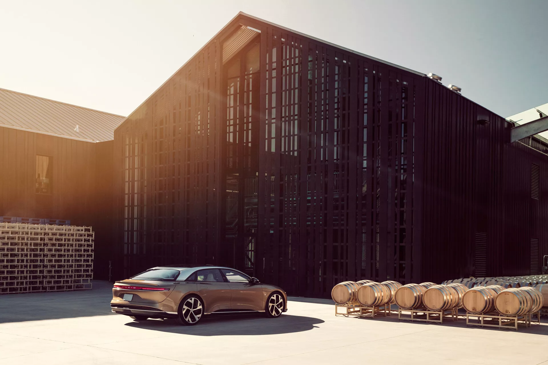 Bild av en 2022 Lucid Air sedan i en industriell miljö, förmodligen ett destilleri eller bryggeri, som visar bilens eleganta aerodynamiska linjer och sportiga design. Den moderna sidan av passagerarsidan framhäver de mångekrade legeringshjulen, LED-belysningsfunktioner och den sluttande baksidan av det lyxiga, högteknologiska fordonet mot en bakgrund av träfat.