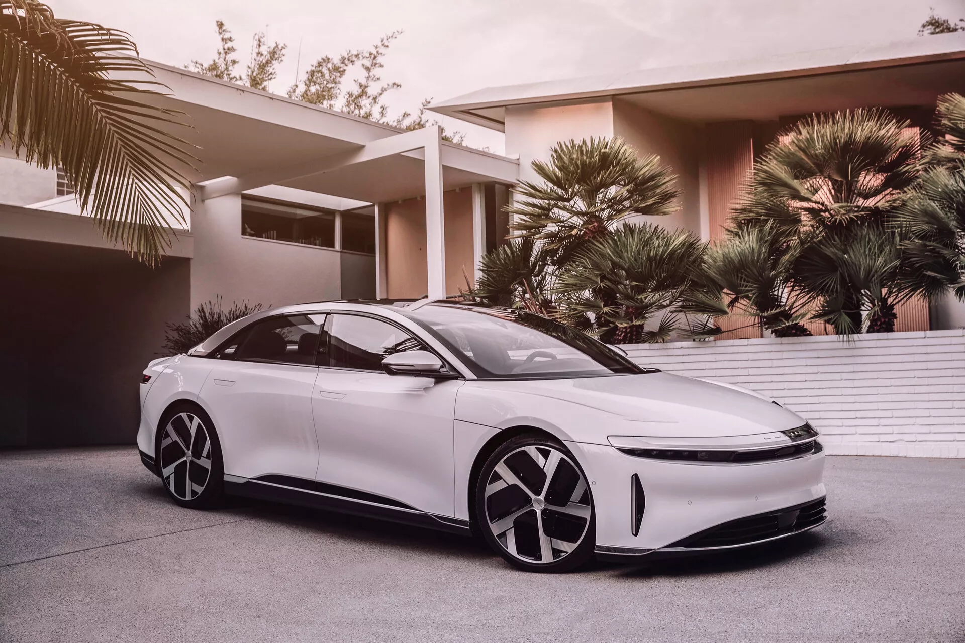 Bilden visar en först generations 2022 Lucid Air, en modern, elegant vit sedan med låg, bred hållning och en kupéliknande taklinje. Bilden är taget från en låg vinkel, vilket framhäver bilens aerodynamiska design, stora distinkta hjul, LED-strålkastare och strömlinjeformade sidospeglar, som antyder avancerad förarassistans-teknologi. Den minimalistiska frontgrillen antyder elektrisk eller hybrid drivlineteknologi. Den futuristiska fälgdesignen har aerodynamiska kåpor och bilens utseende förbättras av tonade rutor, ramlösa dörrar och eleganta, infällda dörrhandtag. En partiell vy av ett panoramasoltak antyder en lyxig, luftigt kupé. Bakgrunden visar ett lyxigt bostadsområde med palmer, vilket tyder på att bilen är riktad mot förmögna konsumenter.