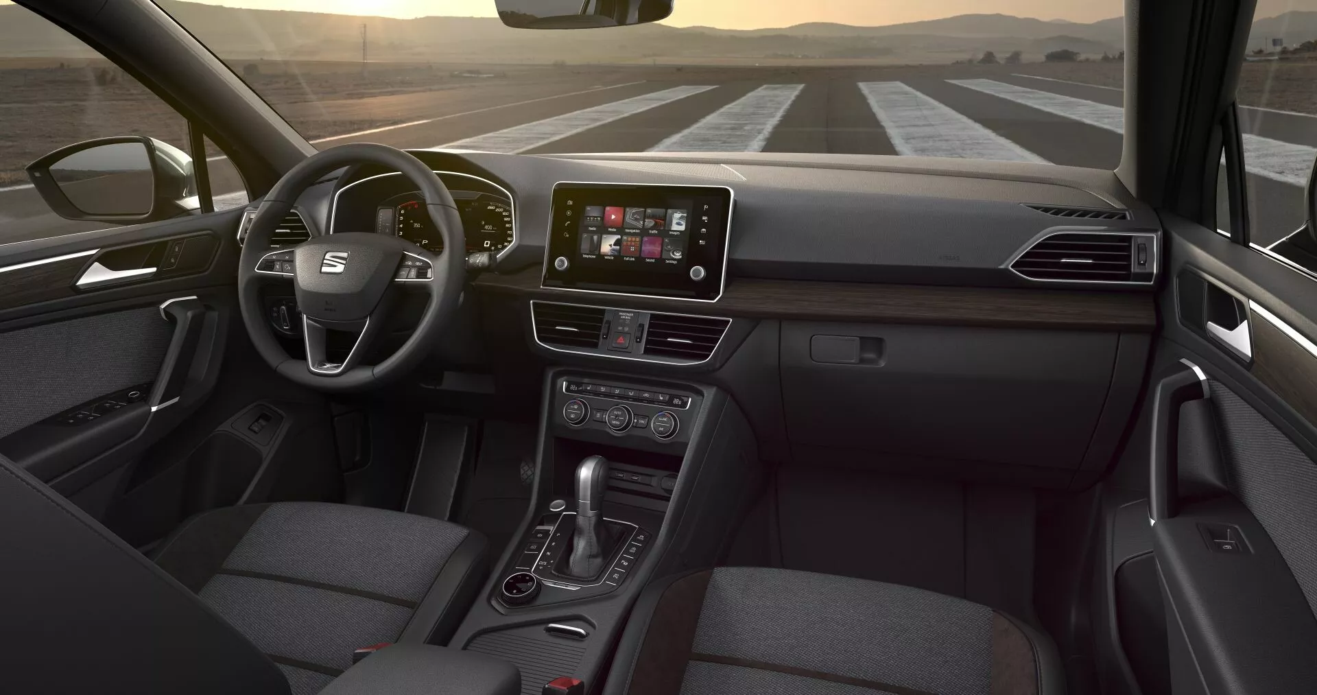Interiörperspektiv av Seat Tarraco 2019 från baksätet, visar multifunktionsratten, den centrala infotainment-pekskärmen, kontrollknappar för klimatanläggningen, växelspak för automatväxellåda och passagerarsäte klätt i tyg, under subtil inre belysning