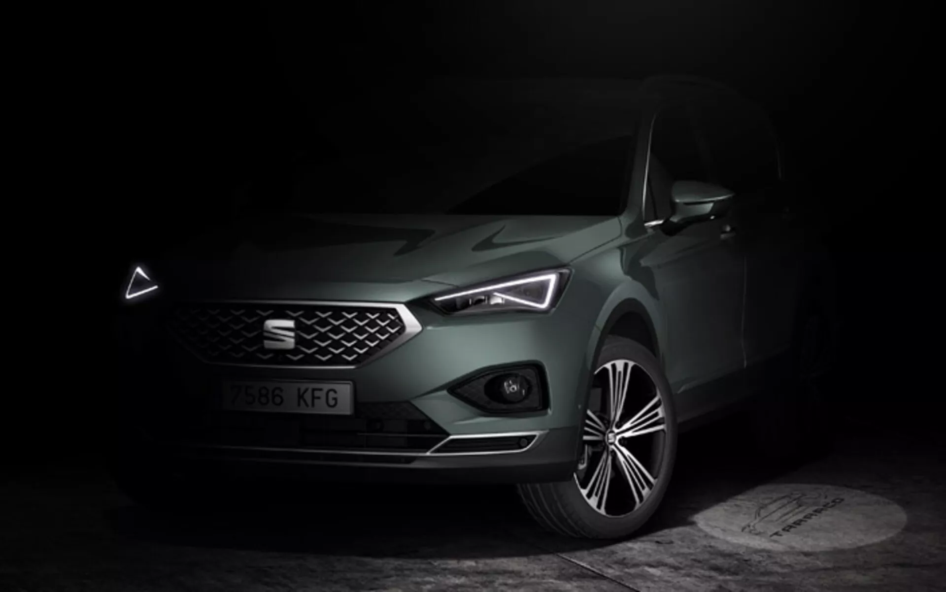 Seat Tarraco 2019 visas i en dunkel, stämningsfull miljö som betonar dess snygga front, geometriska grill, LED-strålkastare med distinkta dagljuslampor och skulpterad motorhuv, tillsammans med en detaljerad vy av legeringshjulet, som framhäver bilens lyx och moderna teknologiska egenskaper