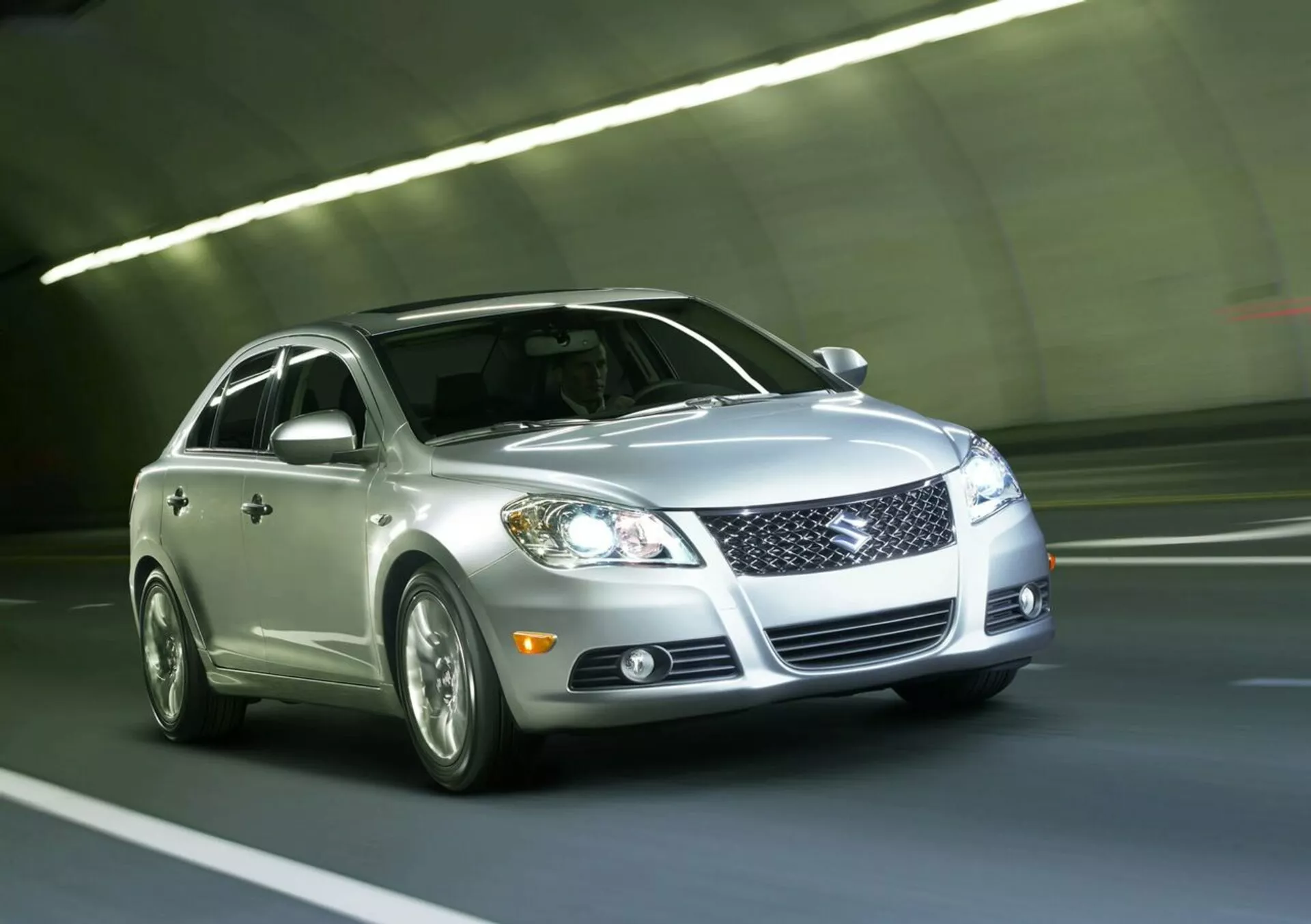 2010 Suzuki Kizashi silver sedan som kör genom en tunnel, med frontgrill, LED-strålkastare, dimljus, lättmetallfälgar och reflekterande lack, sedd från en låg vinkel framifrån och tre fjärdedelar som betonar fordonets dynamiska design och teknik.
