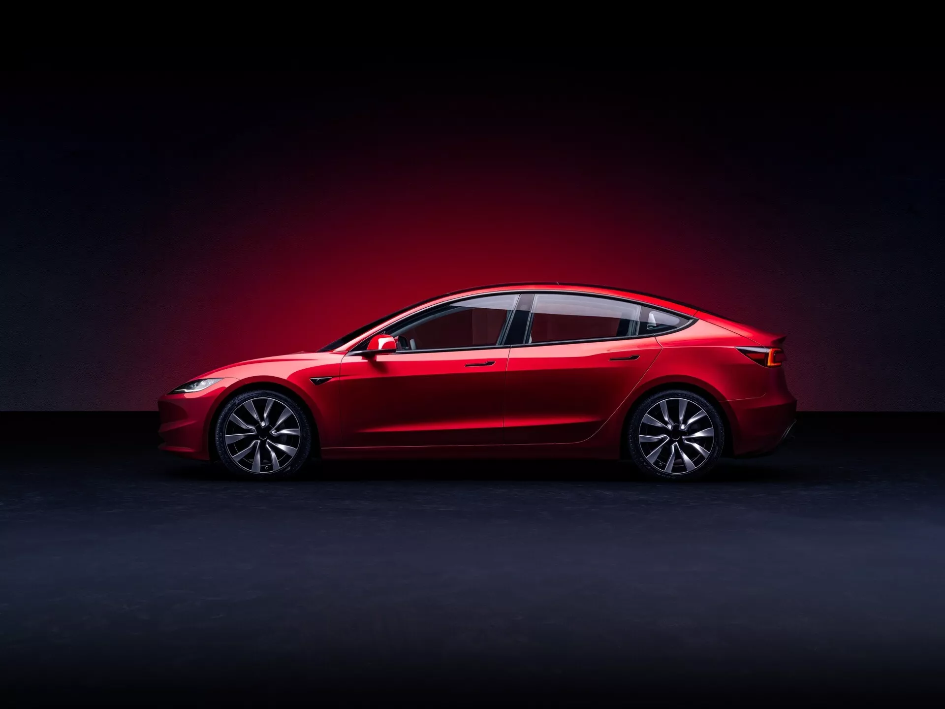 2023 Tesla Model 3 (1:a generationens ansiktslyft) visas från sidoprofil, vilket framhäver dess slanka design, fullängds synlighet inklusive fram- och bakdäck, sidofönster och speglar, med fokus på de aerodynamiska linjerna, legeringshjulen och modern teknik funktioner, inställd mot en dramatisk ljussättning för att betona den glansiga röda ytan