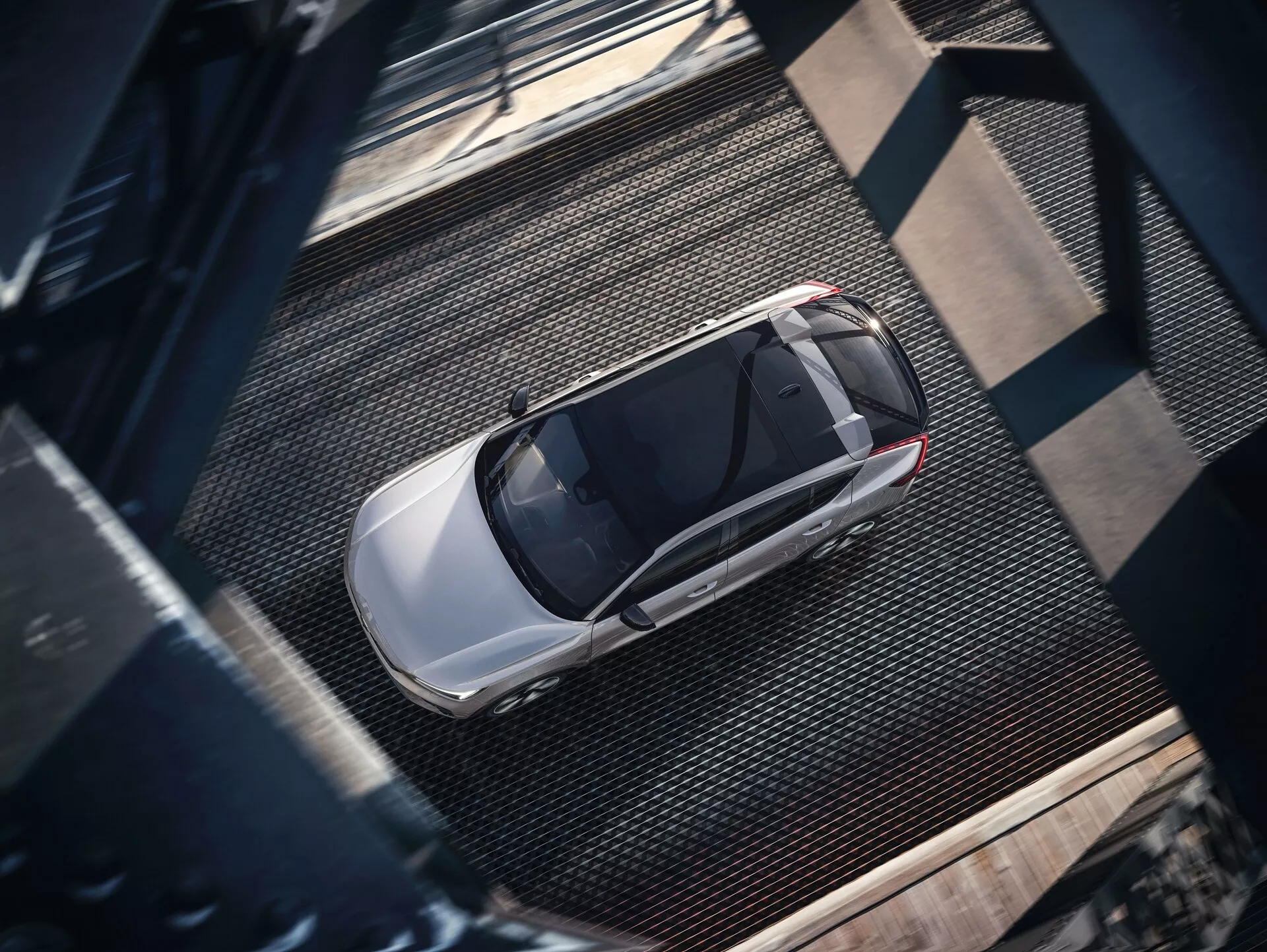 Fågelperspektiv av en Volvo C40 2021 första generationen, fångad från en hög vinkel. Bilden visar en komplett överblick av den eleganta, moderna sedanen med en ljus, metallisk och glansig finish, troligen silver eller ljusgrå. Synliga detaljer inkluderar taket, huven, bagageluckan, fönstren och delar av dörrarna, med ett framträdande soltak som antyder avancerad ventilation. Bilen står parkerad på en texturerad, industriell yta med gallerliknande metalltexturer och mörka nitade balkar, som kastar skuggor som indikerar sideljus, troligen på morgonen eller sen eftermiddag.