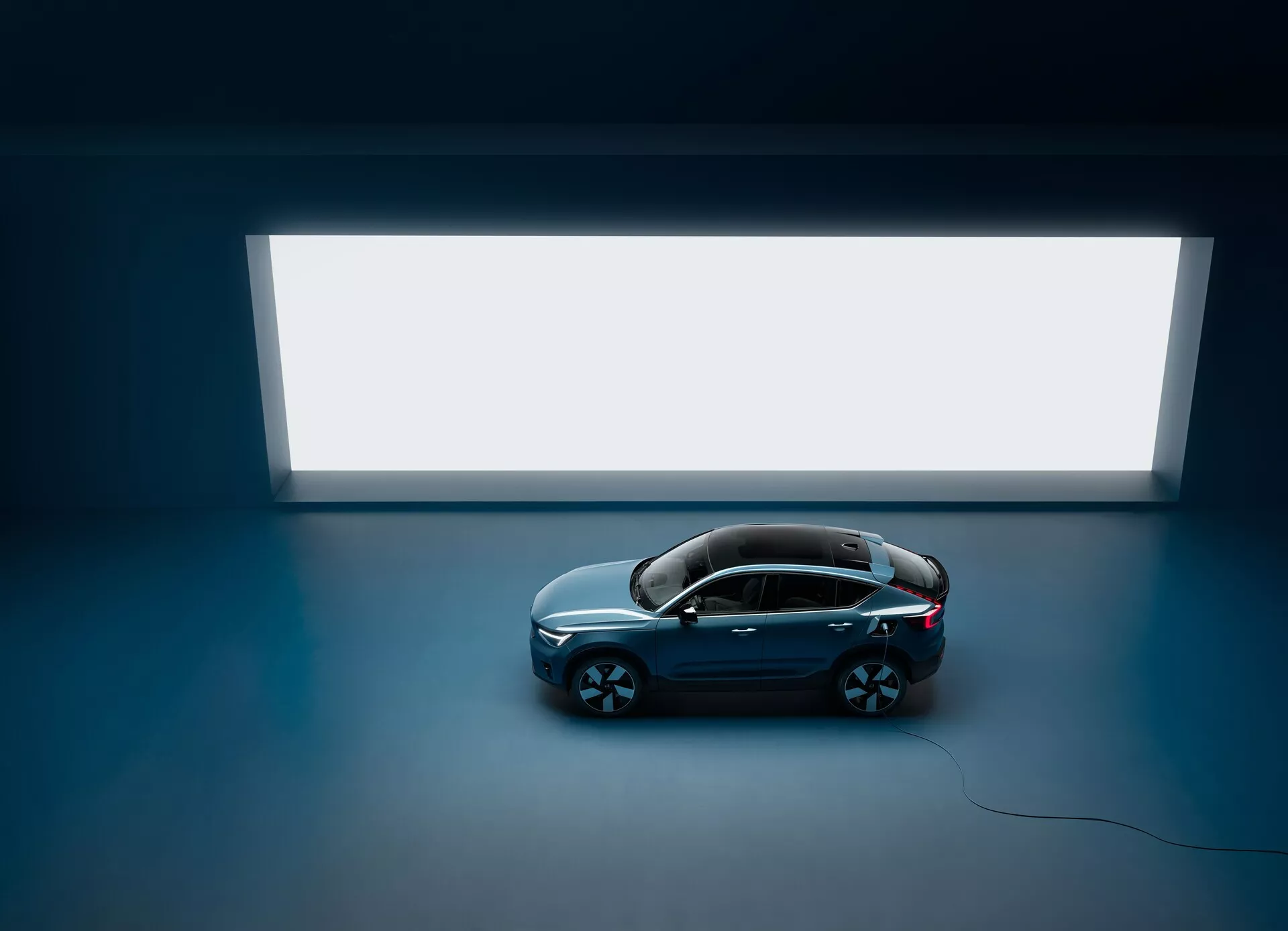Högvinklad bild av en Volvo C40 2021 sedan med fyra dörrar och panoramatak, sedd ovanifrån mot ett mörkt golv med en stor, ljus rektangulär ljuskälla i bakgrunden, som visar en ren och minimalistisk design med en glänsande mörkfärgad kaross och stora flerspokrade fälgar, som framhäver dess lyxiga och moderna estetik.