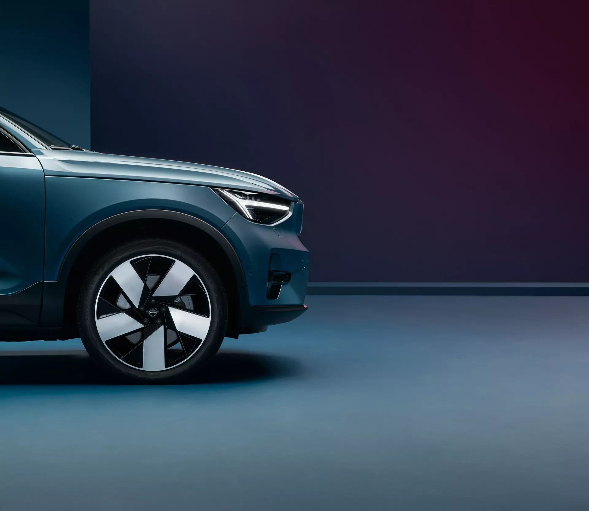 Lågsvinkelbild av 2021 Volvo C40 (första generationen), fokuserad på den främre kvarten från framhjulet till mitten av motorhuven. Höjdpunkterna inkluderar modern design av strålkastarna, flerarmad lättmetallfälg, metallisk glansig lack och aerodynamiska linjer. Den tvåtoniga bakgrunden med mjuk belysning framhäver fordonets premiumfinish och kraftfulla hållning.