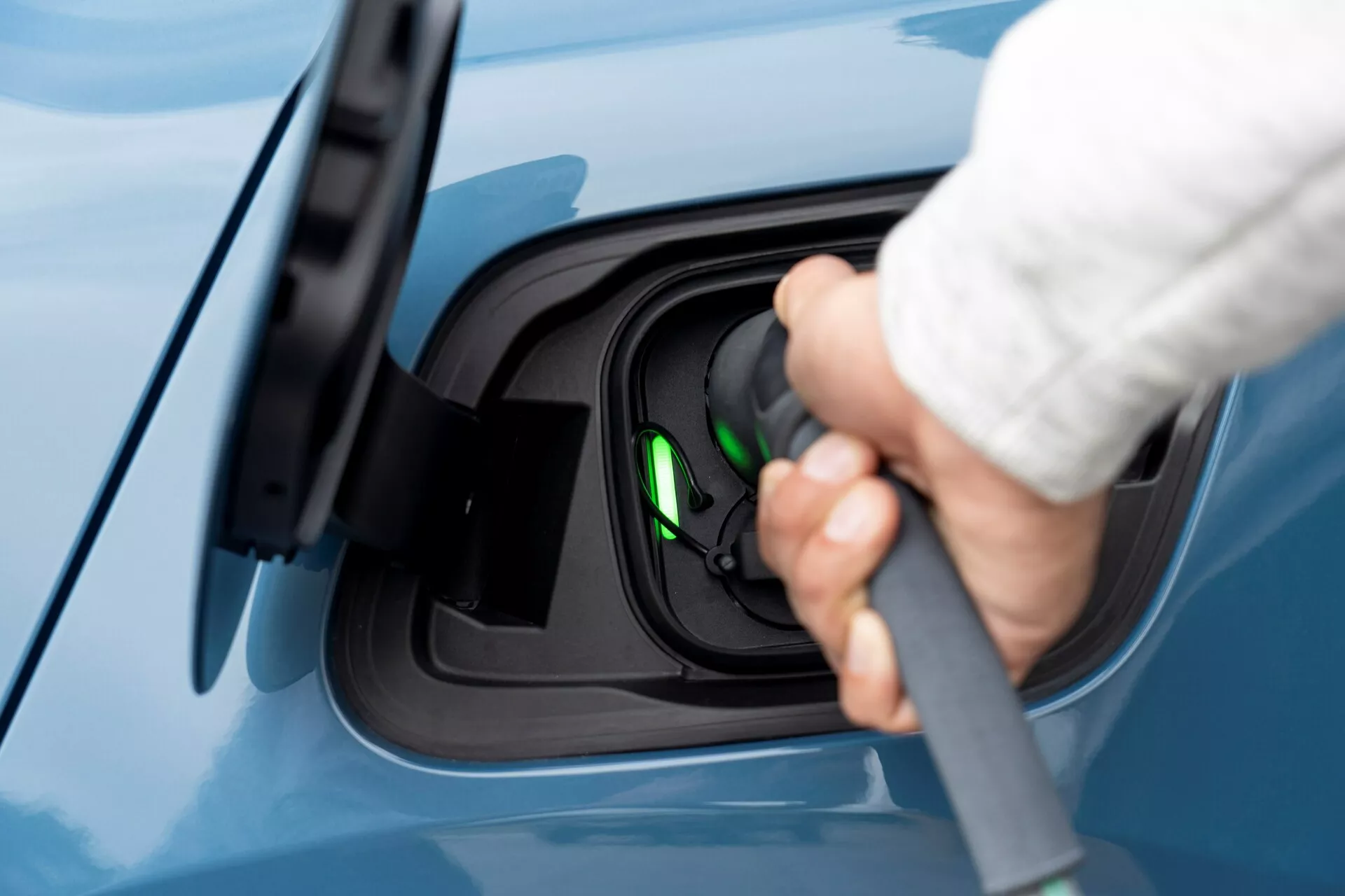 Närbild av laddningsområdet på en 2021 Volvo C40 (första generationen) elbil från sidan, med en hand som ansluter en laddningskabel till porten. Bilden fokuserar på laddningsprocessen och visar en öppen laddport med troligen en Type 2-anslutning. Ett grönt ljus inuti uttaget indikerar att fordonet är redo att laddas eller laddas för närvarande. Vyn är något förhöjd och visar en oblemad blå lack på bilen, vilket betonar det rena och moderna utseendet på elbilen.