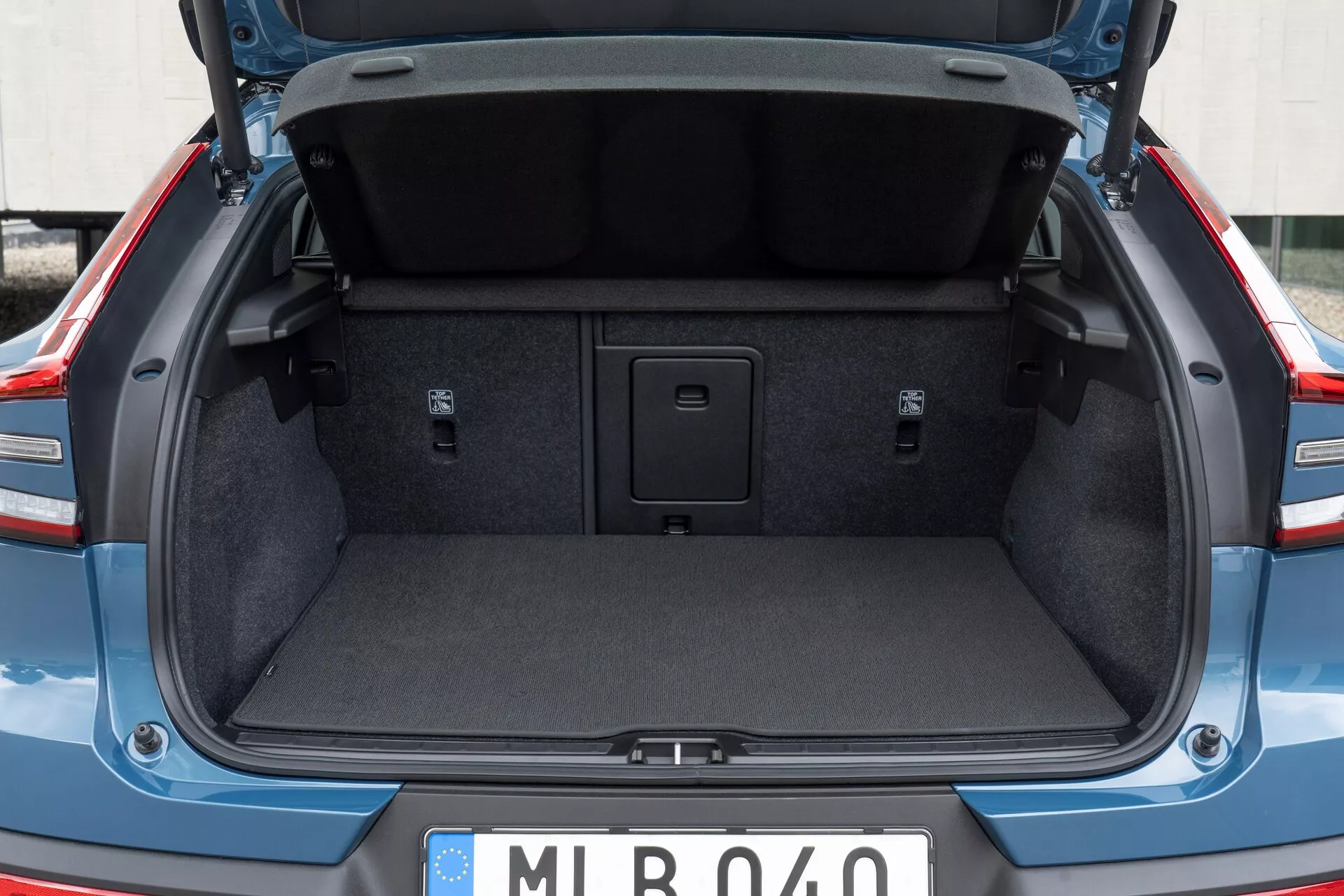 Lastutrymme bak i en modern bil, som visar bagageutrymmet hos en Volvo C40 2021 (första generationen) från ett bakåtperspektiv, något under midjehöjd. Bagageluckan är öppen och lyfter fram den standardmässiga stoppningen på insidan. Bagageutrymmet är tomt, med en mattbeklädd interiör, försänkta sidor och ett handtag i mitten av golvet för åtkomst till ett förvaringsutrymme. Inga lastkrokar eller fästpunkter syns. Den europeiska registreringsskylten antyder en europeisk härkomst.