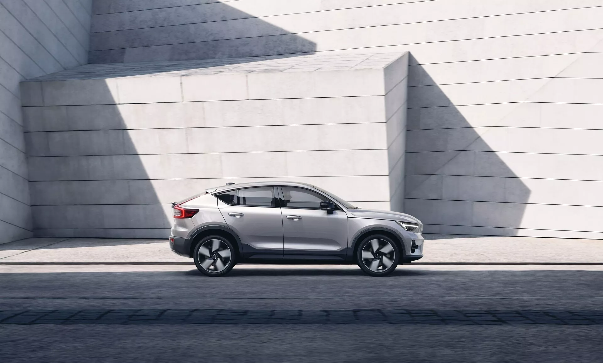 Bild av en modern Volvo C40 2021 SUV parkerad parallellt, som visar vänster sidoprofil från vänster framlykta till vänster baklykta. Vinkeln framhäver markfrigång och hållning på en asfalterad yta. Solljus skapar skuggor på den blanka lackytan. SUV:en har en elegant design med en mjukt sluttande taklinje, distinkta dörrkonturer, tonade rutor och samtida sidospeglar. Fälgar av lättmetall med ett flerspokat mönster och lågprofildäck indikerar prestanda och lyx. Sidokjol, hjulbågsformningar och en karaktärslinje som löper genom dörrhandtagen är synliga och antyder robusthet. Avsaknaden av synligt avgasrör antyder en möjlig elektrisk eller hybriddrivlina. De minimalistiska betongstrukturerna i bakgrunden kontrasterar med bilens raffinerade kurvor och betonar dess urbana och designfokuserade attraktionskraft.
