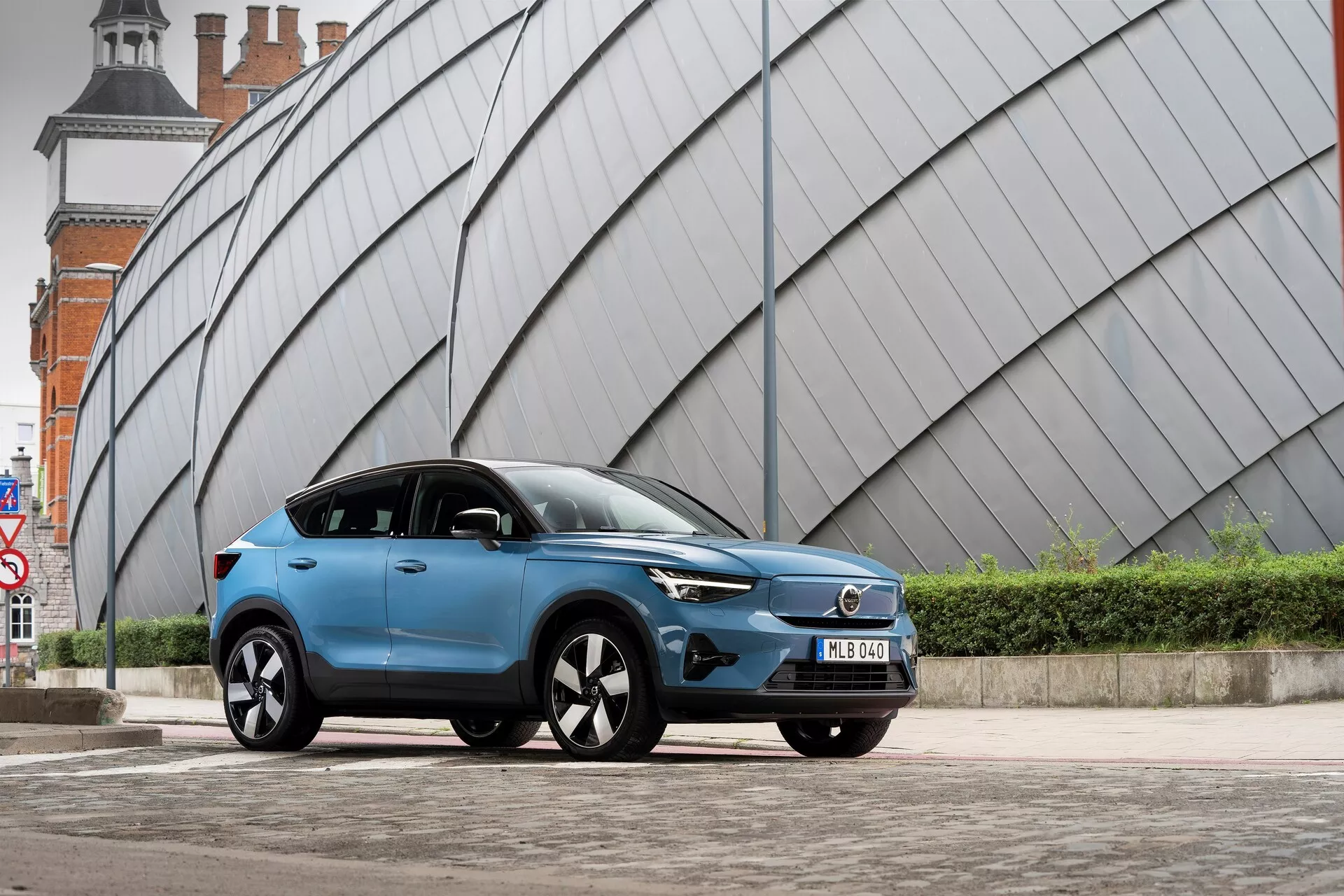 En modern blå Volvo C40 2021 SUV parkerad på ett asfalterat urbant område med en vinklad, metallisk struktur i bakgrunden. Lågvinkelbilden från vänster framsida visar fordonets främre grill, LED-strålkastare, aerodynamisk profil, flerspoked lättmetallfälgar, sidospeglar med integrerade blinkers, matt eller satinlackad färg, tonade rutor, främre registreringsskylt och skulpterade sidolinjer på karossen.