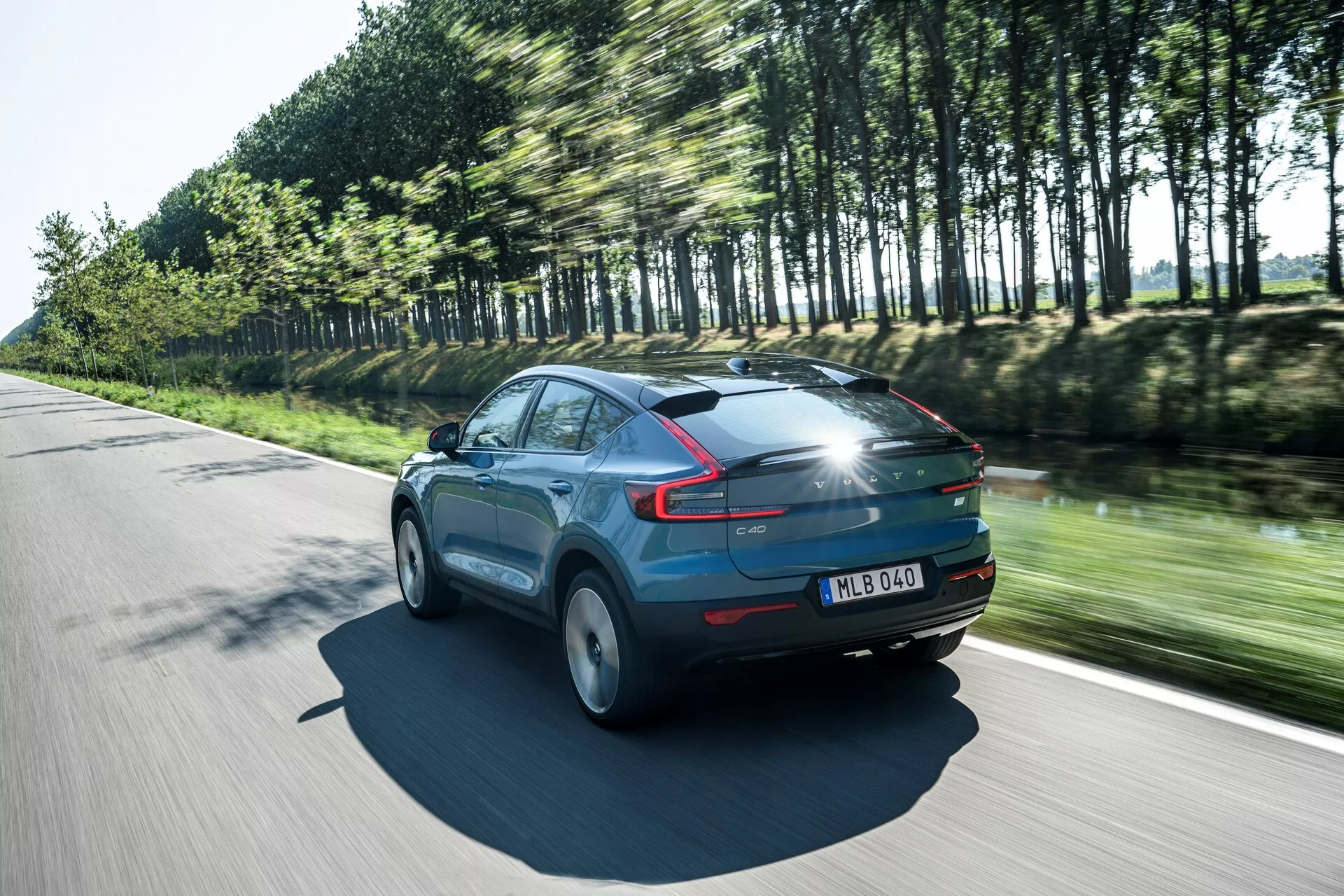 Dynamisk bild av Volvo C40 2021 (1:a generationen) i rörelse på en solig dag, trekvarts bakifrån som visar bilens släta profil och aerodynamik. Bilen, målad i en glansig blå färg, har utmärkande LED-bakljus, rörelseoskärpa på hjulen och träd som speglar dess hastighet. Noterbara detaljer inkluderar ett panoramatak, en bakspoiler och registreringsskylt på bakluckan, vilket framhäver moderna designelement och avancerad belysningsteknik.